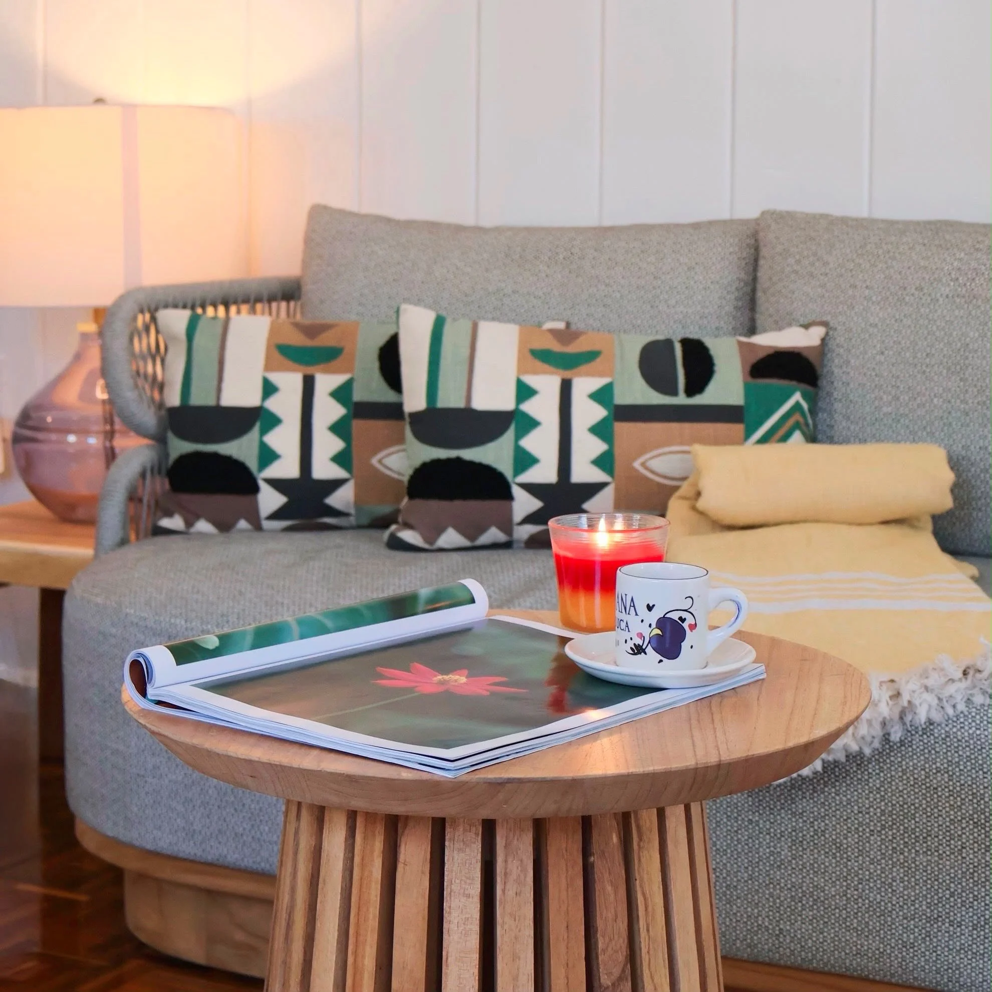 Living room with a grey cushioned sofa, decorative pillows with bold geometric patterns, a wooden side table with magazines, and lit table lamps, against a white paneled wall.