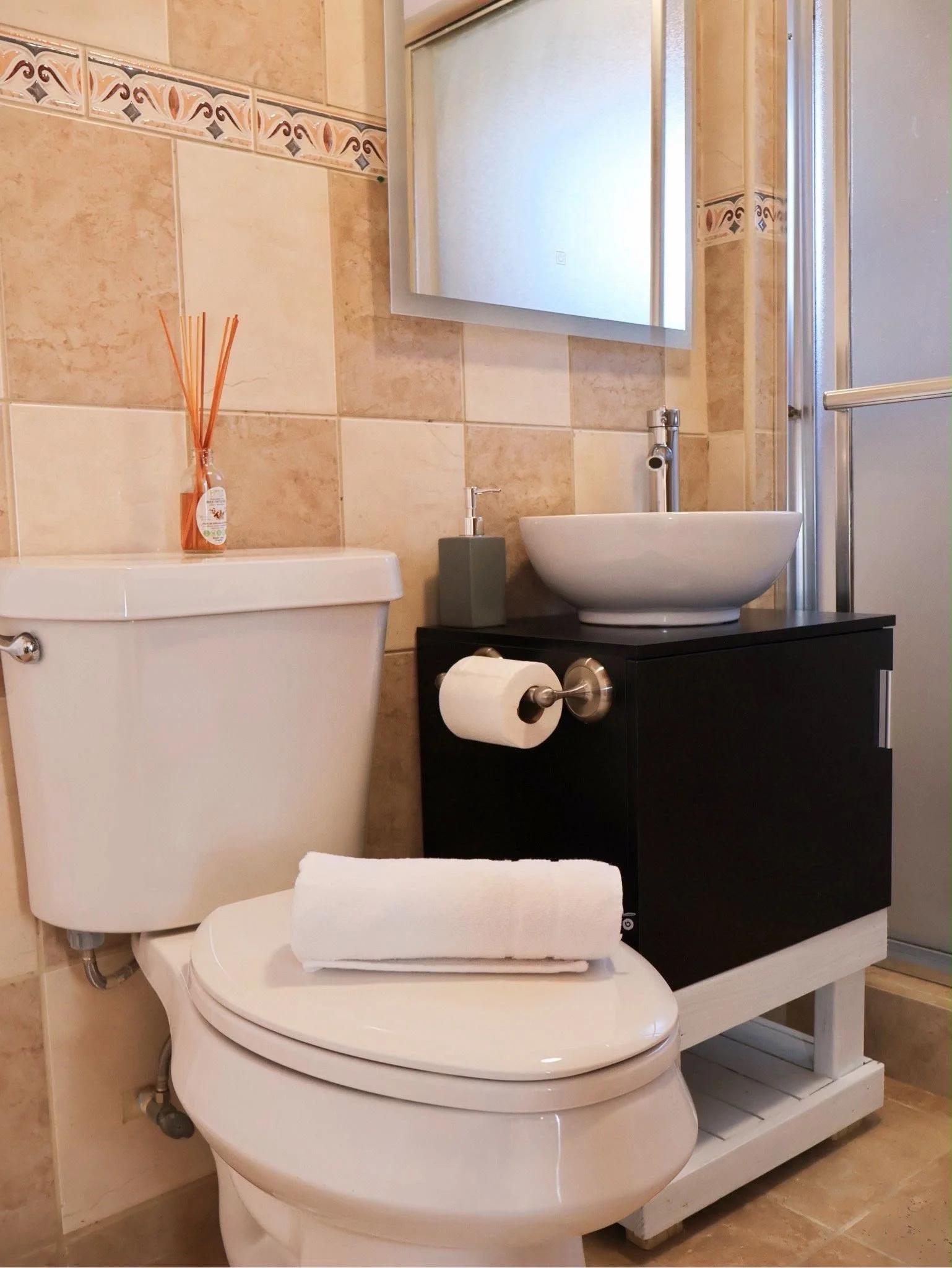 A modern bathroom with beige tiled walls, a toilet with a closed lid, and a set of neatly folded white towels on the toilet seat. There is a small black vanity with a round vessel sink, a soap dispenser, a white mirror, and a glass shower door visibl