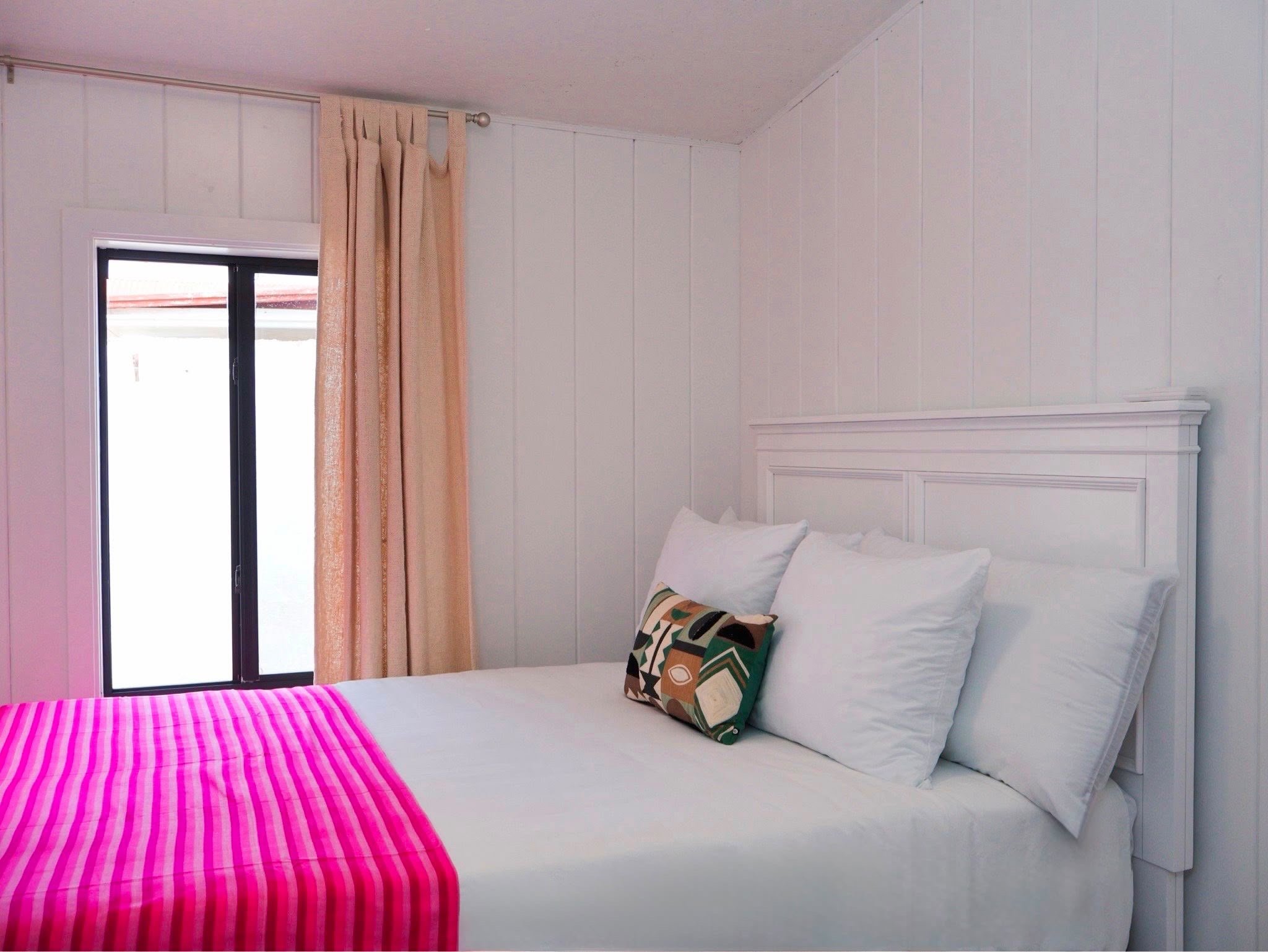 A white bed with pillows, a pink striped blanket at the foot, and a colorful pillow, next to a window with pink curtains in a room with white panel walls and a white headboard.