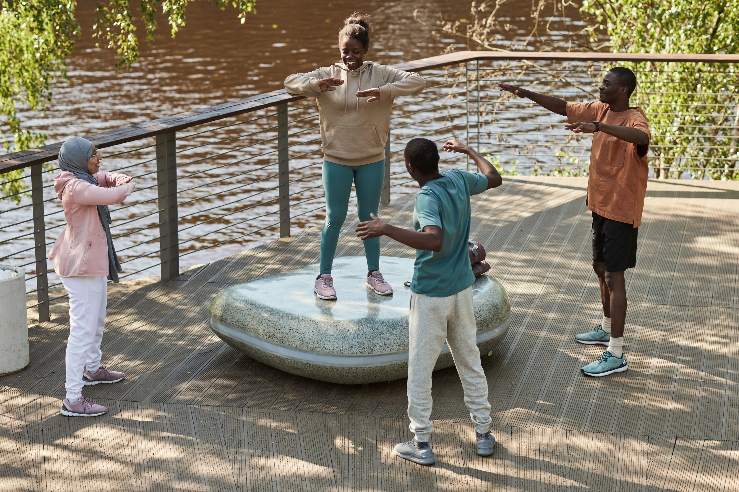 Group of five diverse people participating in outdoor yoga or stretching exercise near a water body, with some standing on a circular platform and others on the wooden deck, during daytime with sunlight filtering through trees.