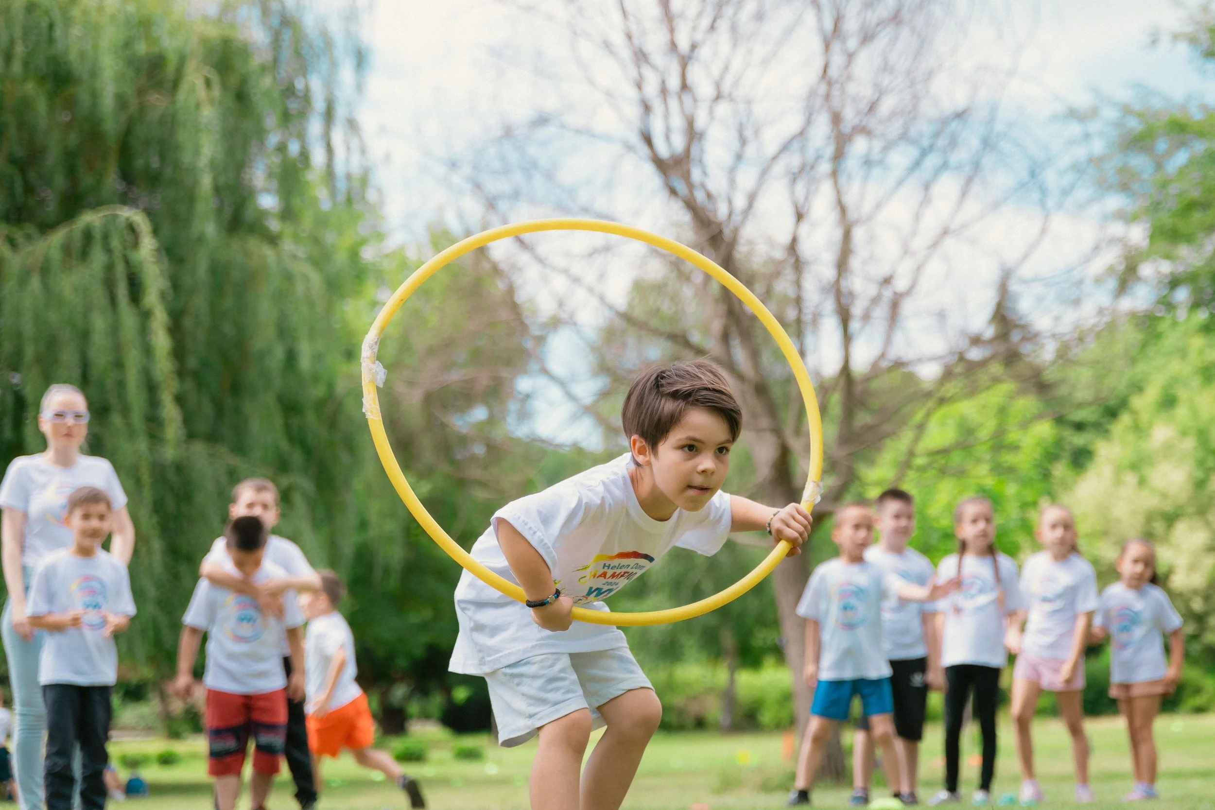 A young boy jumping through a yellow hula hoop held by another person outdoors, with children and adults watching in the background during a daytime outdoor event.