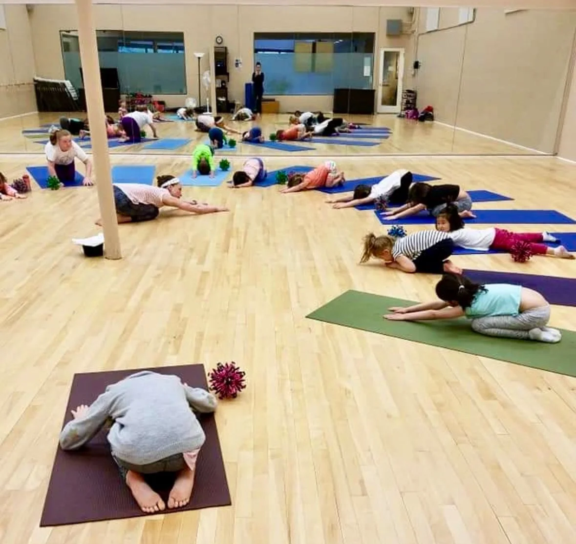 Children participating in a yoga class in a studio, doing a forward bend pose on their mats, with a teacher present and a mirror reflecting the room.