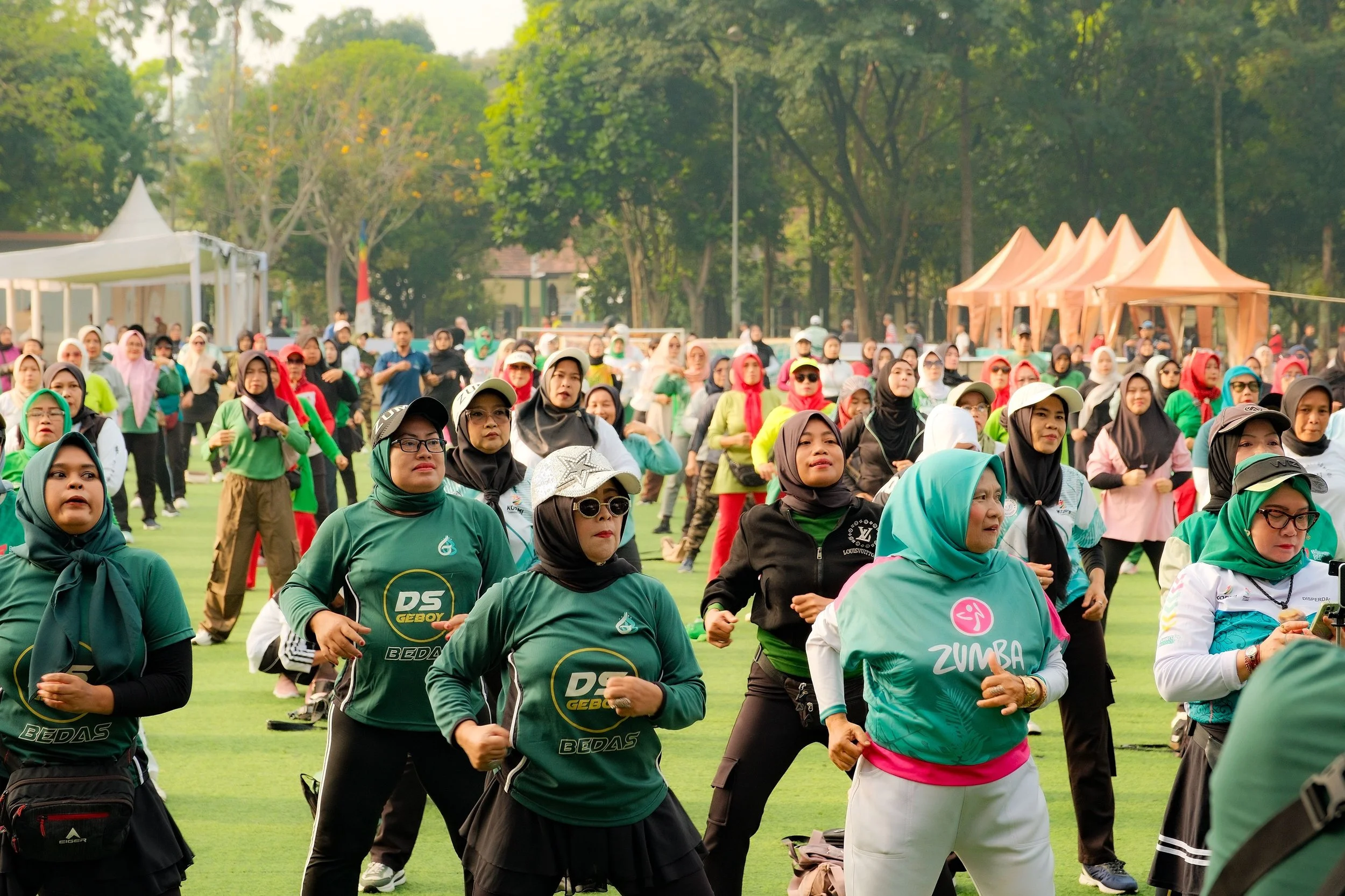 A large group of diverse women participating in a group exercise session outdoors on a grassy field, with trees and tents in the background, during daytime.