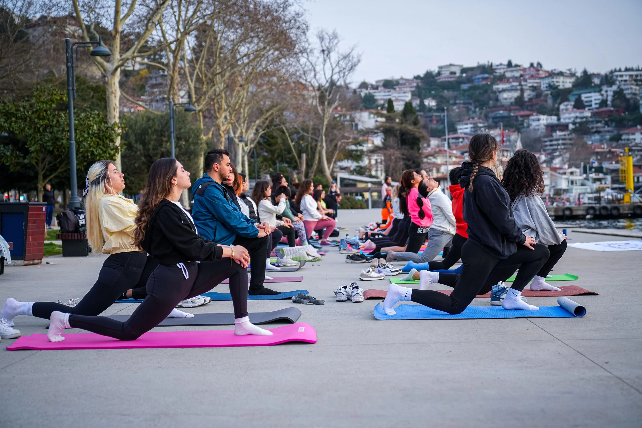 People participating in an outdoor yoga class or meditation session, kneeling on yoga mats by a waterfront with city buildings and trees in the background.