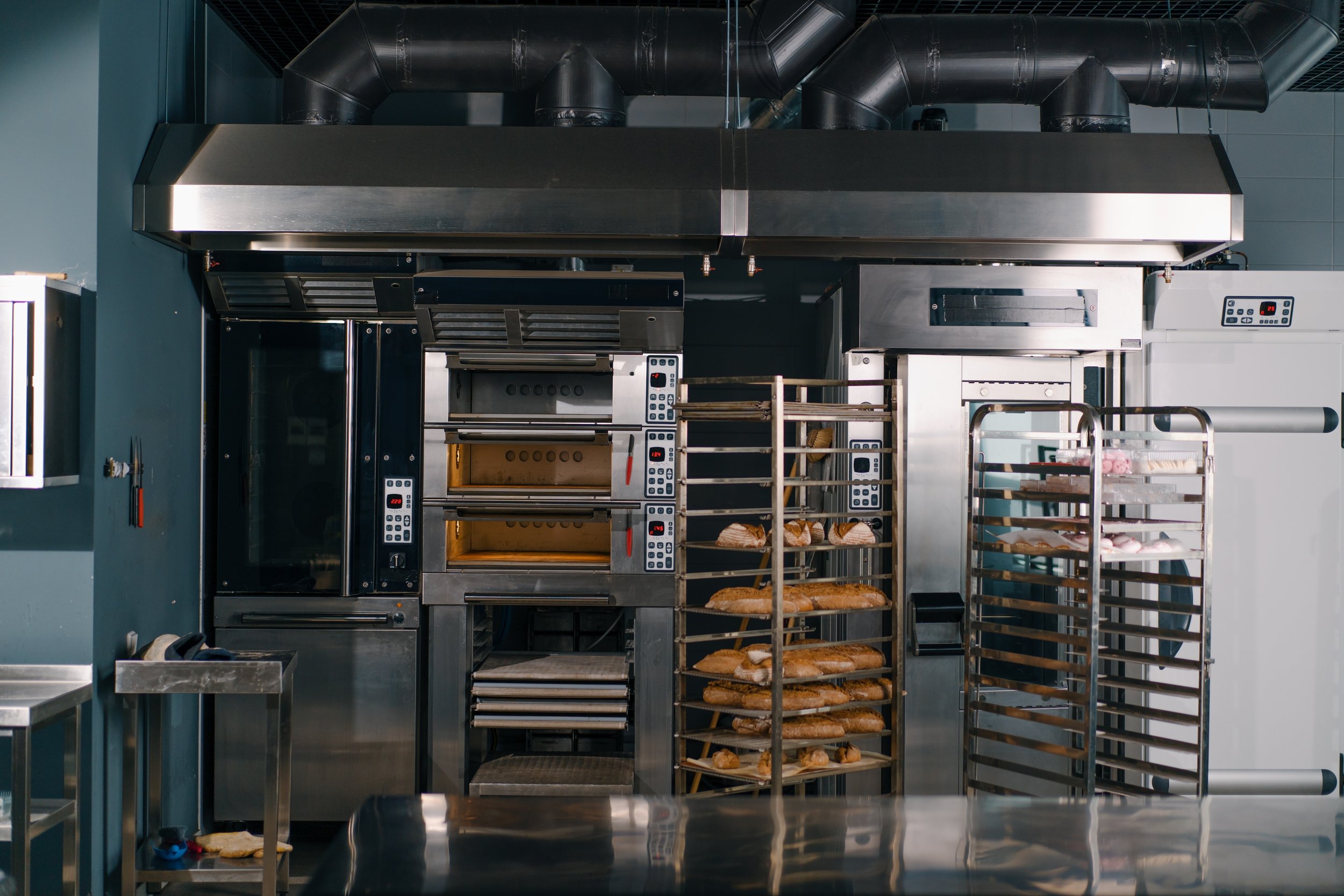 Commercial kitchen with industrial ovens, stainless steel racks of baked bread and pastries, and large ventilation ductwork overhead.