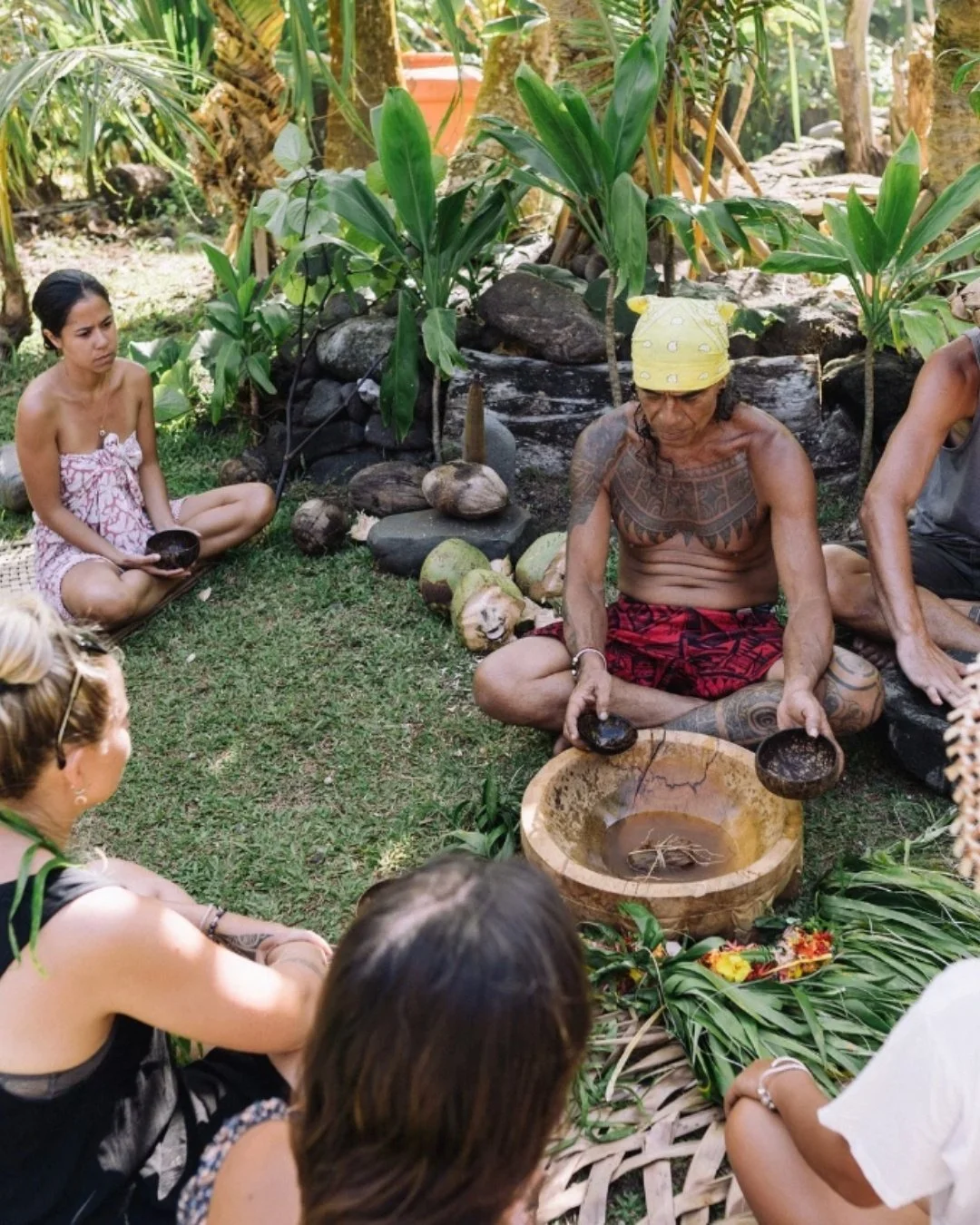 Un groupe de personnes assises en cercle dans un jardin tropical, un homme tatoué en pagne rouge tient une coupe en bois, au centre un bol en bois contenant de l'eau, entouré de feuilles et de fleurs.