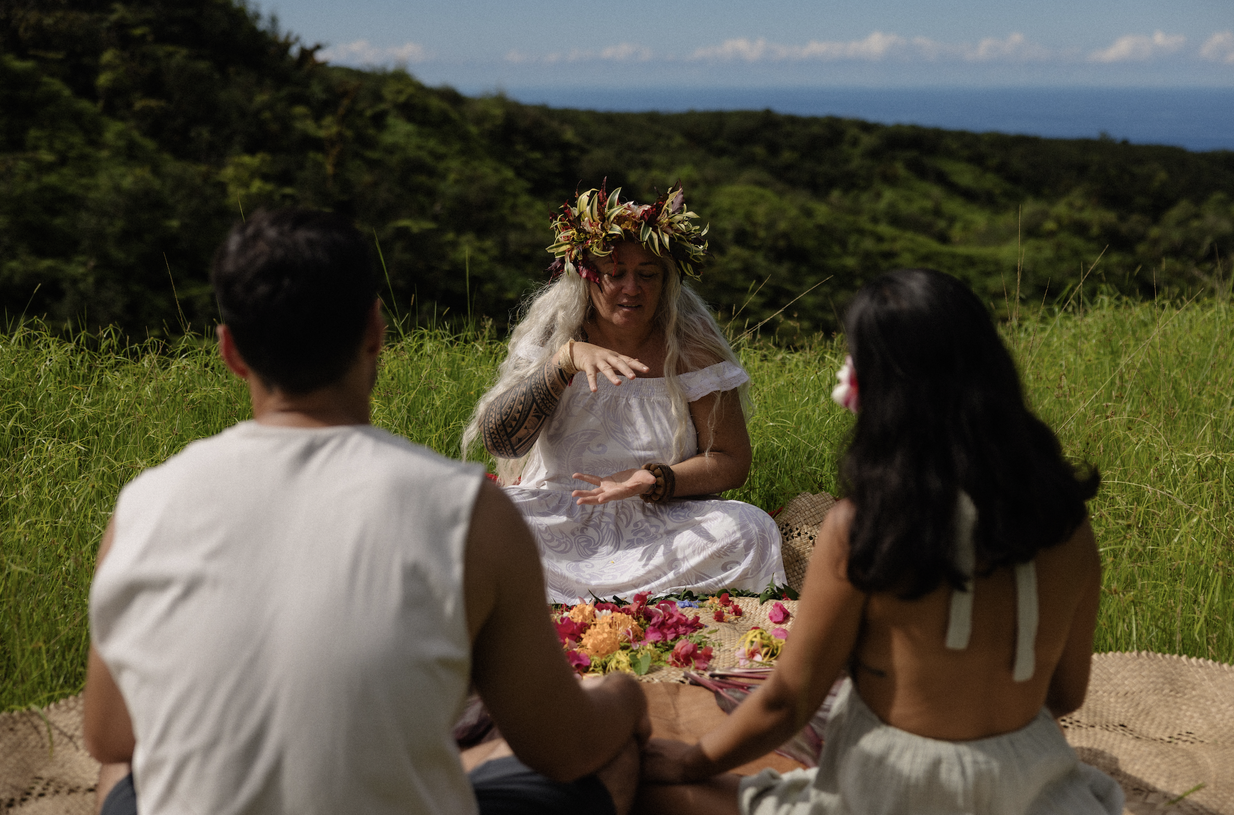Un homme et une femme en tenue traditionnelle maorie, avec des décorations corporelles, rasés de près, prêts à déposer des fleurs blanches près d'une vieille pierre dans un jardin vert, en train de faire un rituel.