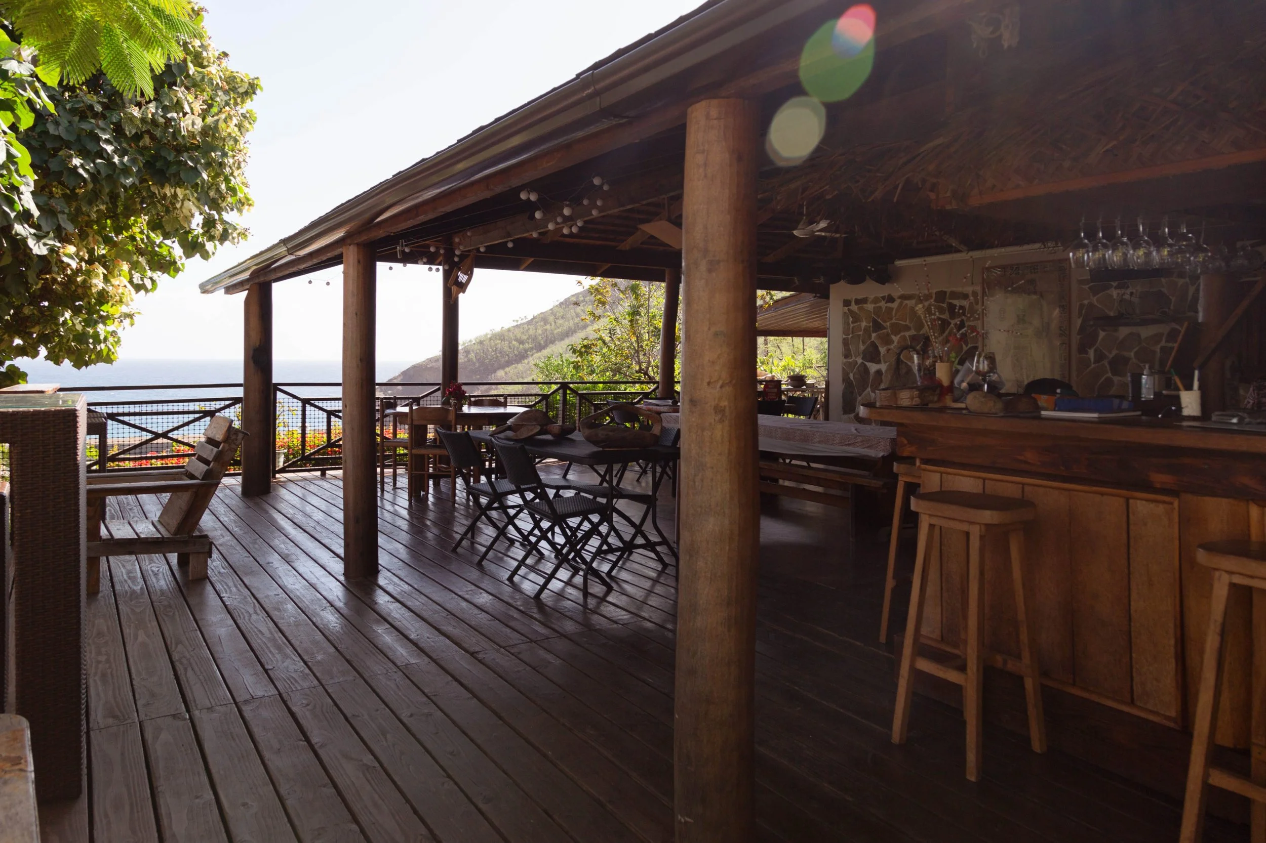 Un espace extérieur en bois avec des tables et des chaises, entouré de végétation et offrant une vue sur la mer et une montagne au loin