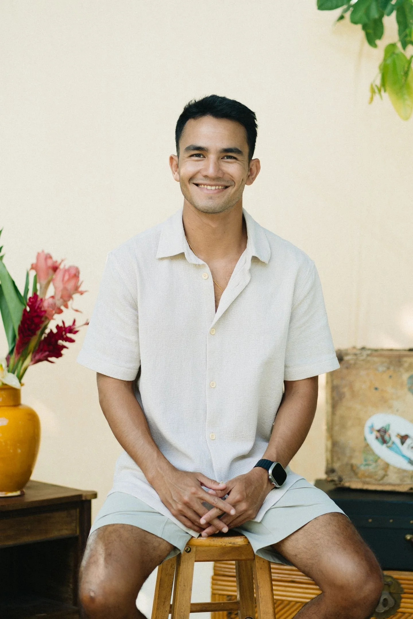 Un homme assis sur un tabouret en bois, portant une chemise blanche à manches courtes, des shorts beige, une montre noire, et un bracelet doré. Il sourit, tenant ses mains jointes sur ses jambes. À gauche, une grande vase jaune avec des fleurs roses et rouges. En arrière-plan, un mur beige, une plante verte en haut à droite, et une table en bois avec une pièce en céramique peinturée.