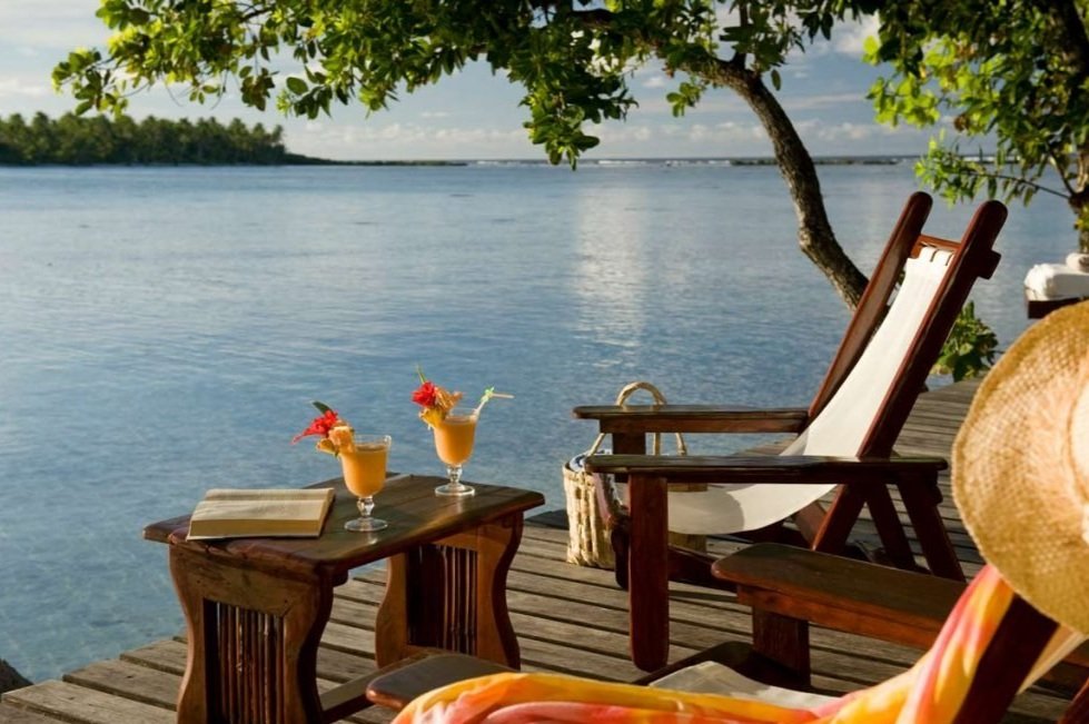 Une scène de détente sur un pont en bois au bord de l'eau avec deux fauteuils en bois, des cocktails avec des fleurs, une tablette, et un chapeau, entouré d'arbres et de nature.