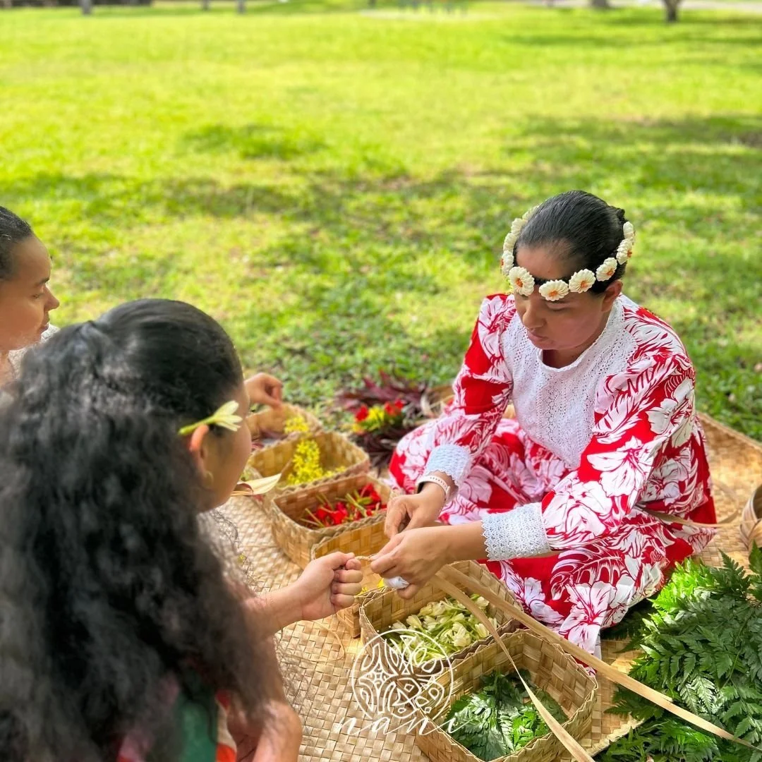 Une femme vêtue d'une robe rouge et blanche avec une couronne de fleurs blanches, assise en tailleur sous un arbre, échangeant des fleurs avec deux autres femmes. Expérience authentique en polynésie,
