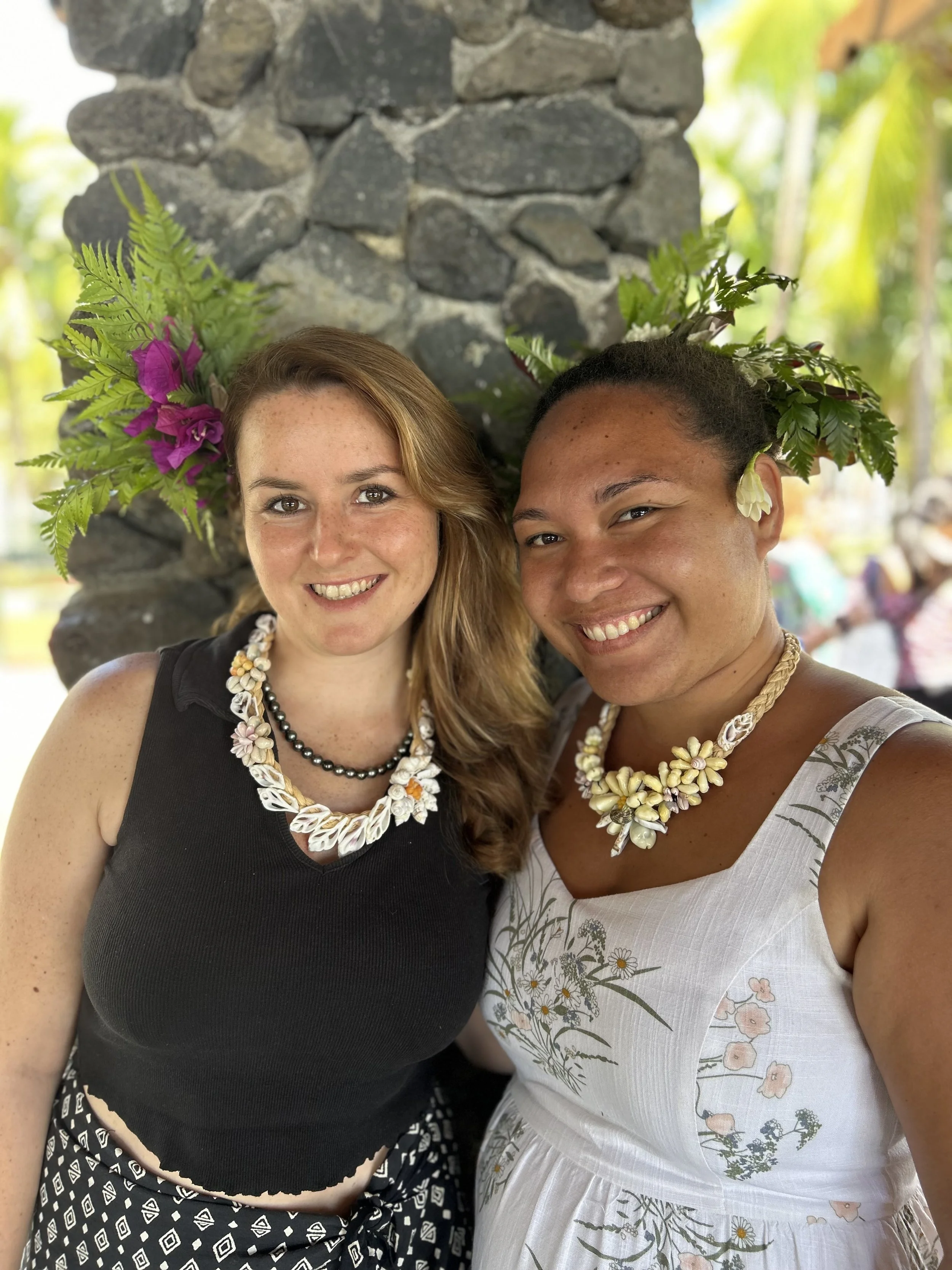 Deux femmes souriantes portant des colliers de coquillages, posant devant un mur en pierre avec des fleurs dans leurs cheveux.