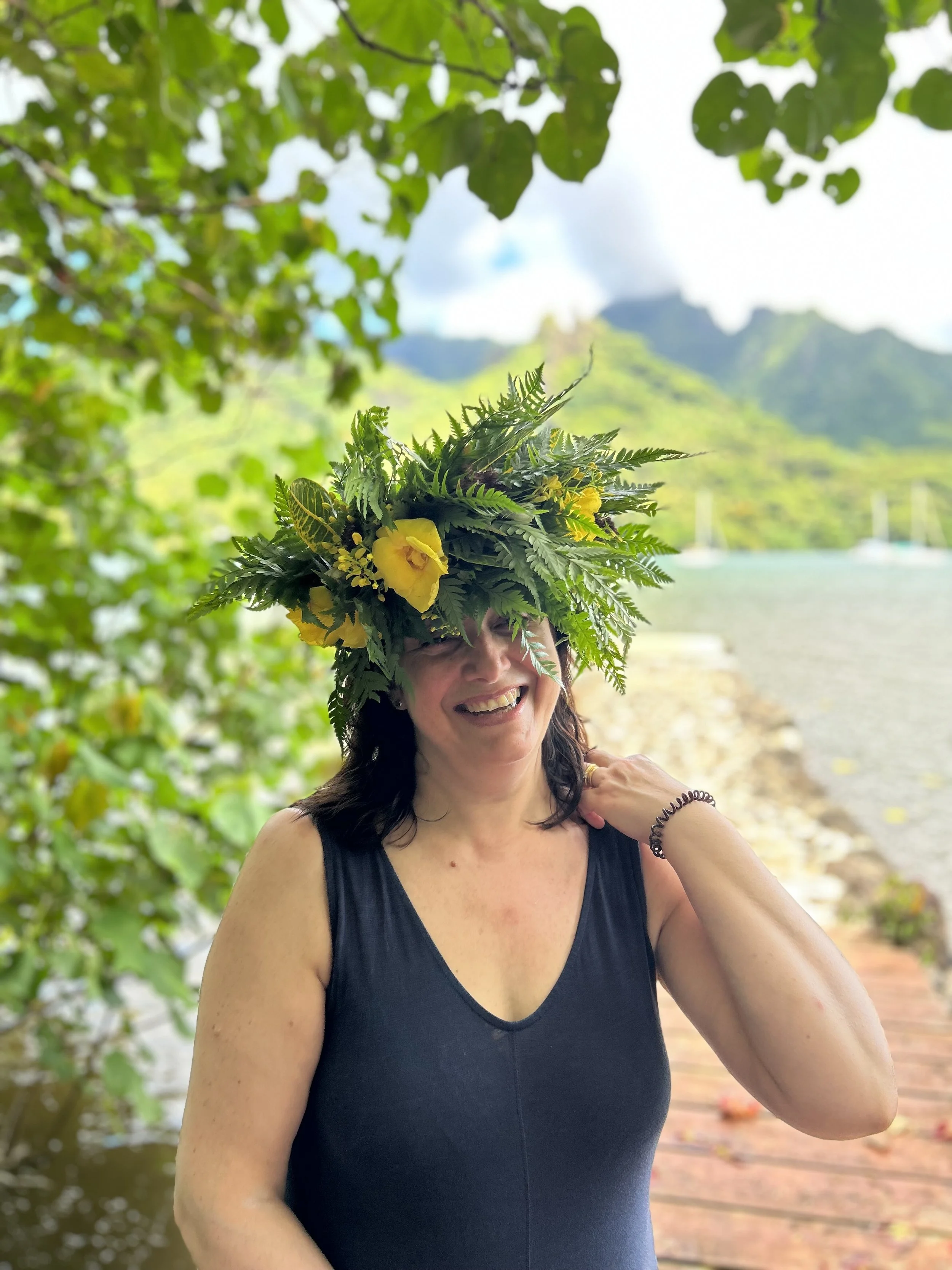 Femme souriante portant un grand chapeau décoré de feuilles et de fleurs jaunes, avec un arrière-plan naturel de montagne, mer et ciel nuageux.