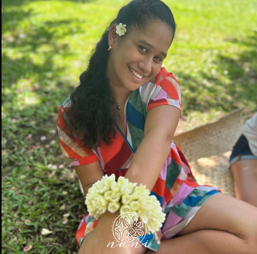 Une femme souriante portant une robe à motifs colorés, avec une fleur dans les cheveux, assise dans un espace vert, tenant un bracelet de fleurs blanches.