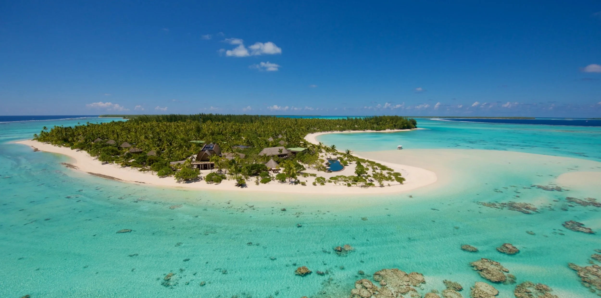 Une île tropicale avec des bâtiments en bois, entourée d'eau turquoise claire et de plages de sable blanc, sous un ciel bleu avec quelques nuages.
