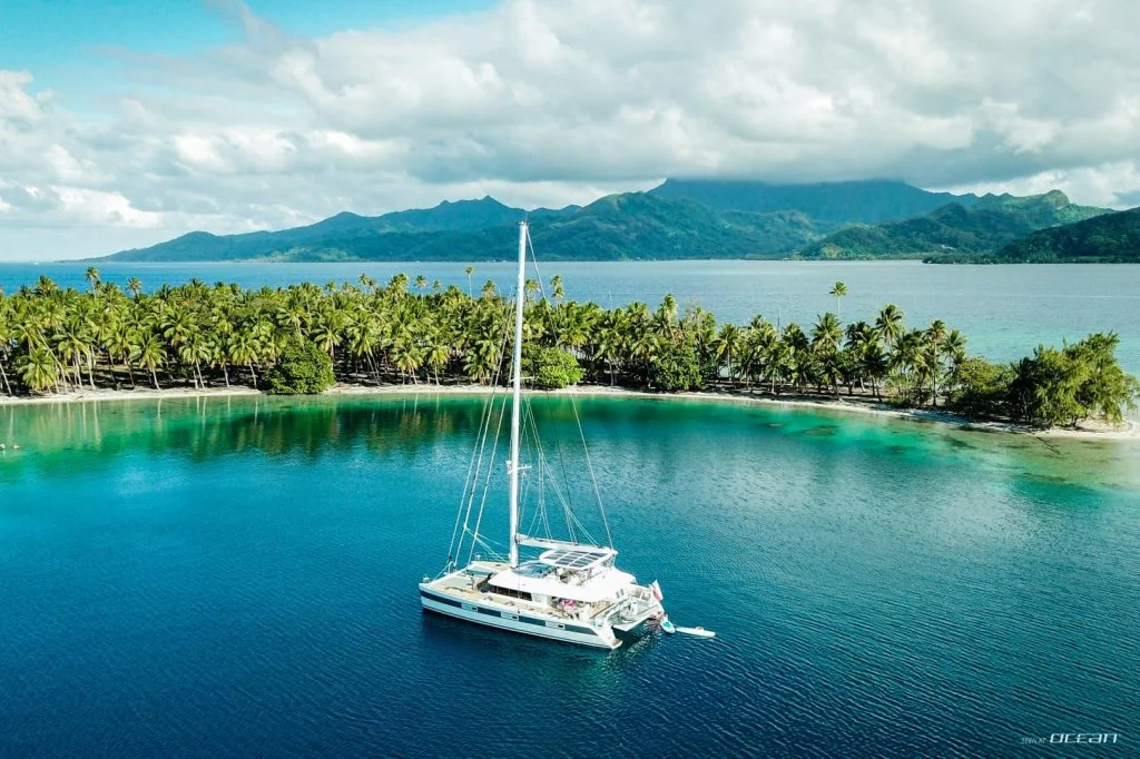 Un yacht blanc est amarré dans une baie turquoise entourée de cocotiers, avec des montagnes à l'arrière-plan et un ciel partiellement nuageux.