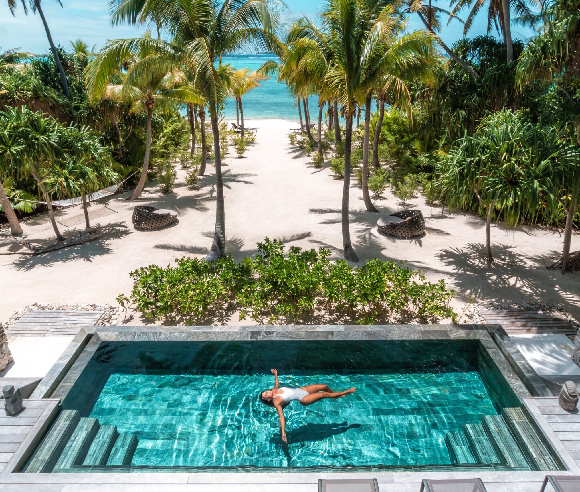Une femme fait un plongeon dans une piscine en face d'une plage bordée de palmiers et de l'océan bleu sous un ciel ensoleillé.