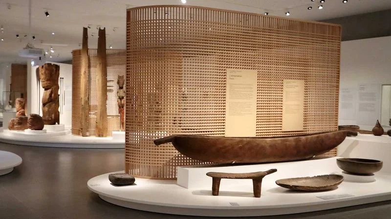 Exposition dans un musée avec des sculptures en bois et des objets en céramique, affichés sur des tables blanches, et une structure en bois perforée au fond.
