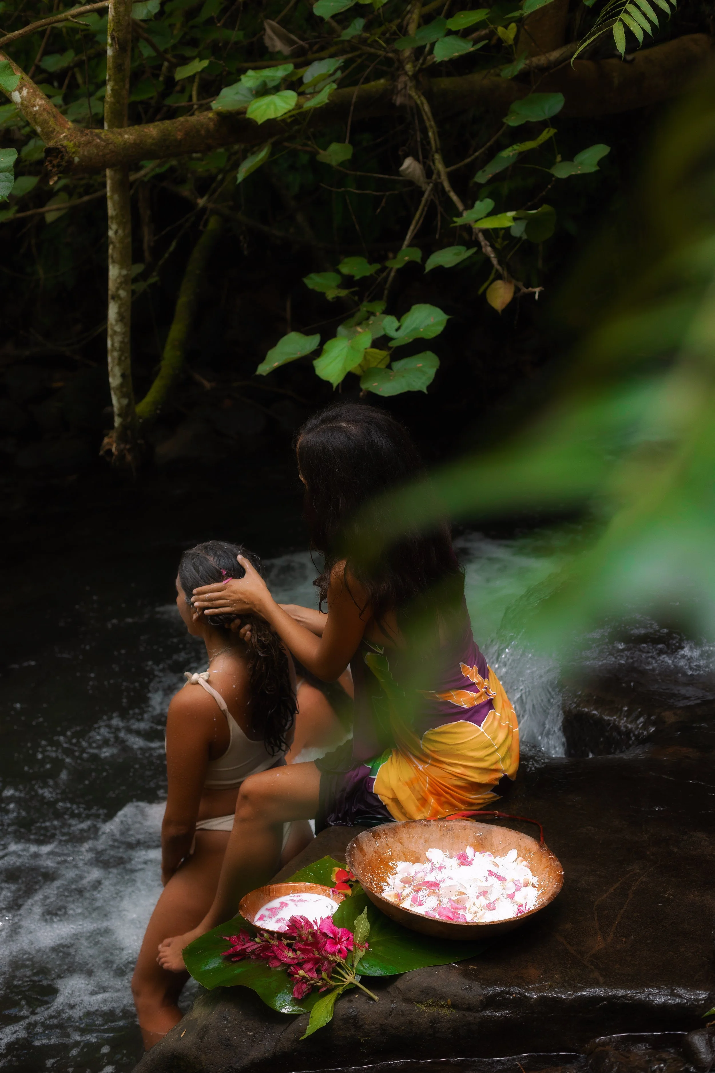 Deux femmes en maillots de bain, une assise dans l'eau et l'autre debout derrière elle, dans une rivière, entourées de végétation, avec des fleurs dans un plat en bois posé sur une pierre devant elles.
