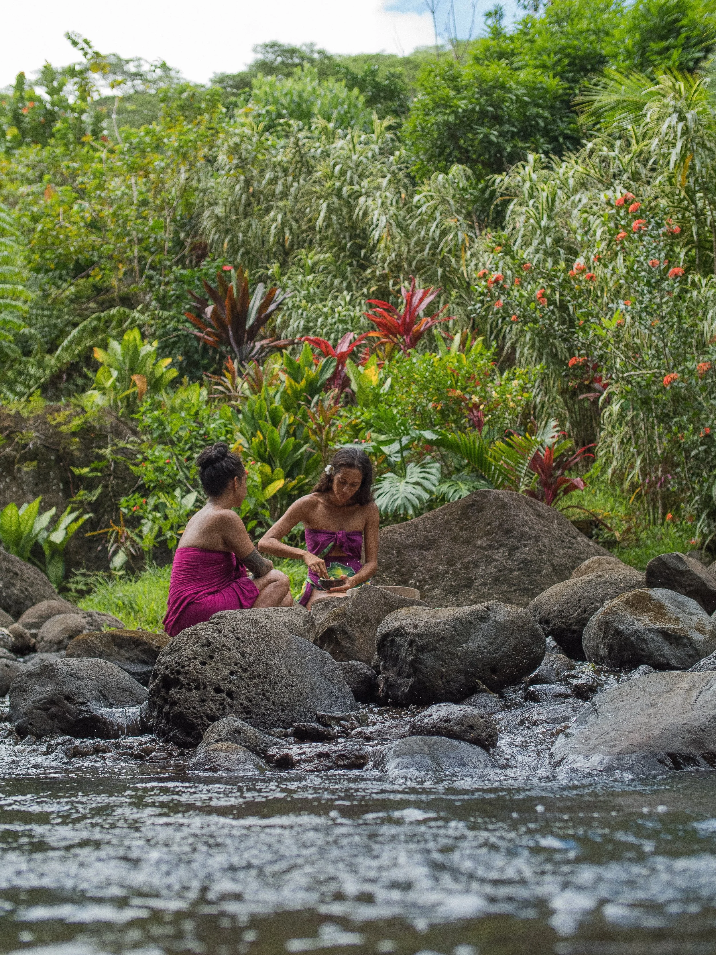 Deux femmes en robes roses assises sur des rochers au bord d'une rivière en forêt tropicale