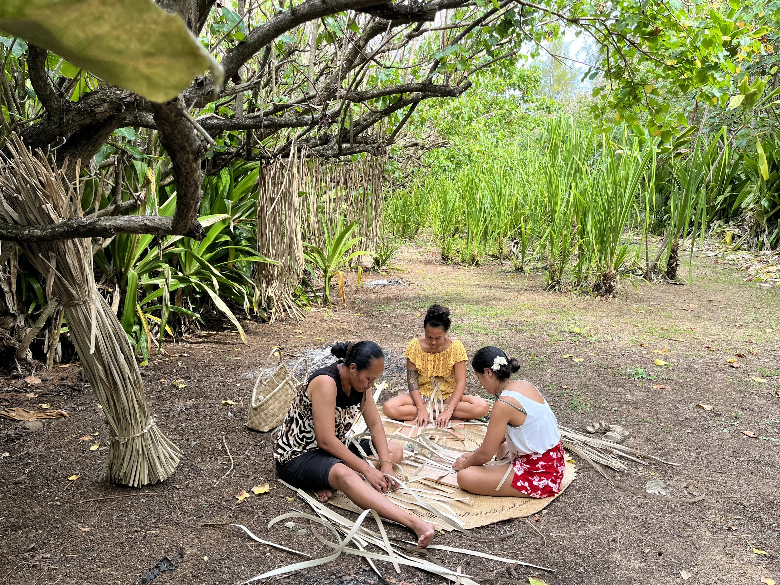 Trois femmes asiatiques en vêtements légers, assises par terre dans une forêt verdoyante, tressent des feuilles de palmier pour fabriquer un objet traditionnel.