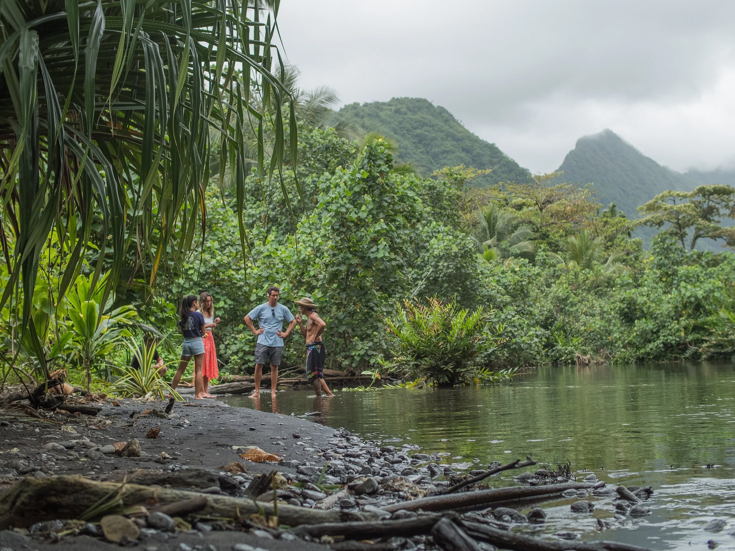 Groupe de personnes discutant au bord d'une rivière entourée d'une jungle verte et montagneuse, sous un ciel nuageux.