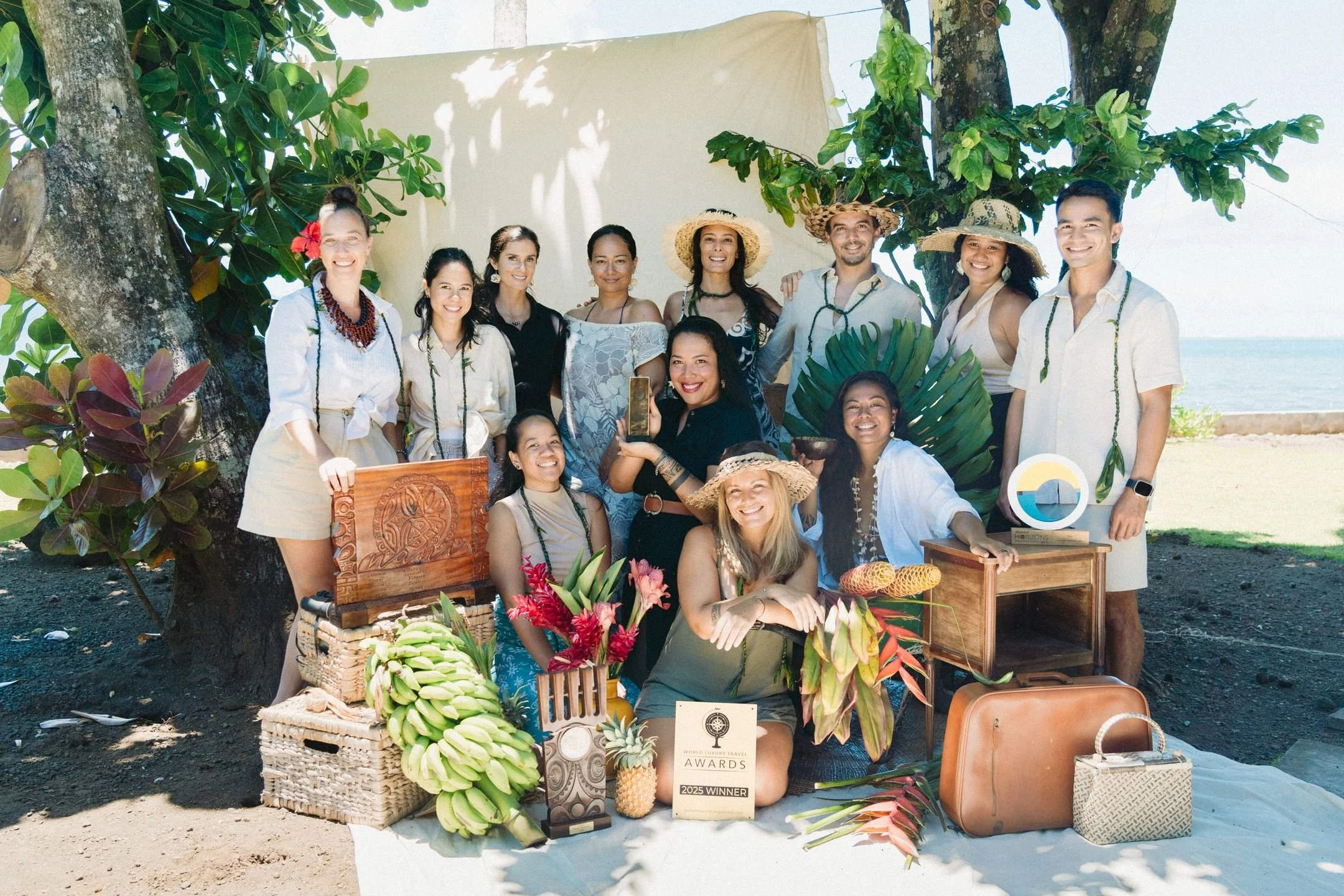 Un groupe de personnes souriantes posant lors d'un événement en plein air, avec des tables et des décorations tropicales, devant une mer ou un océan.