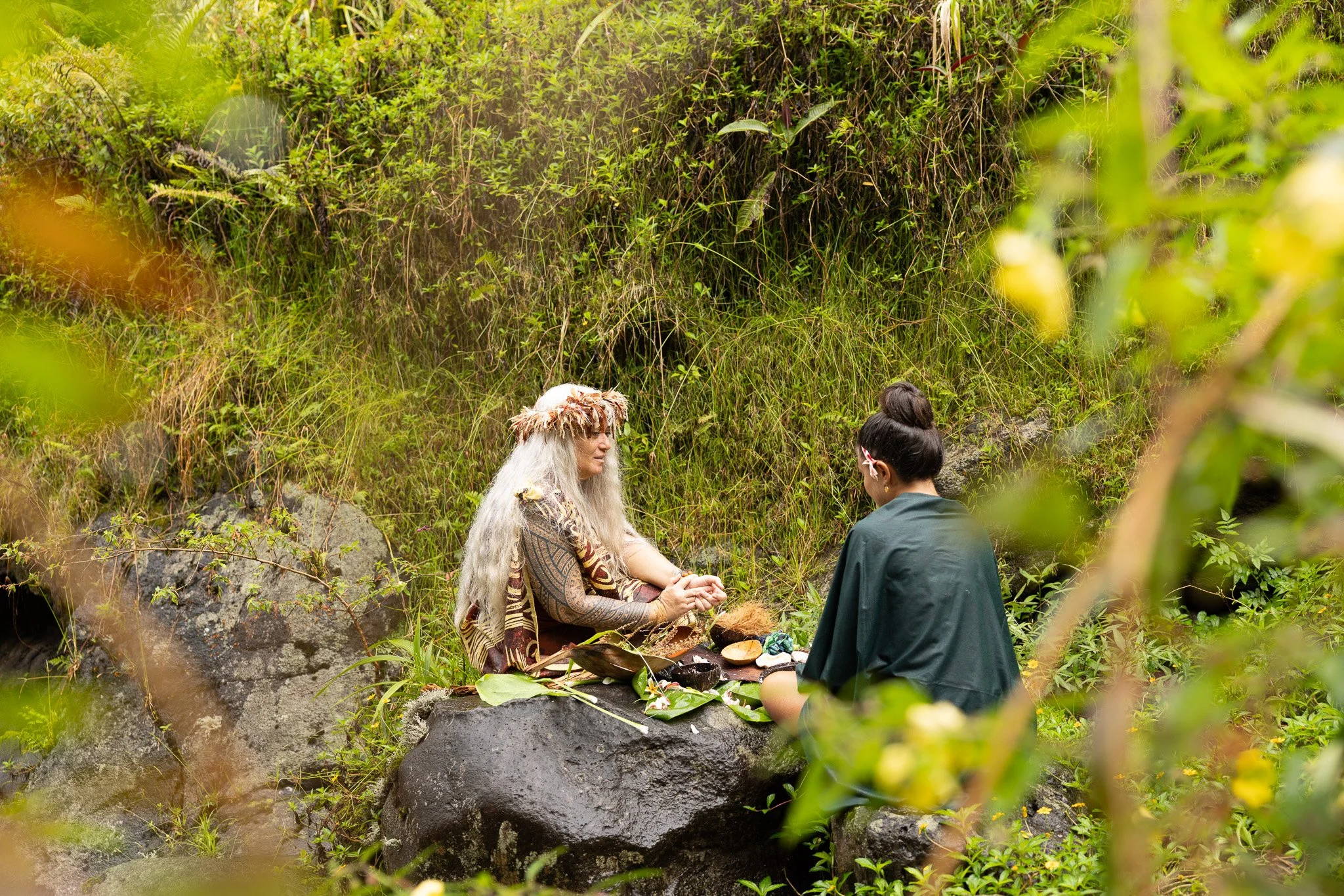 Deux personnes en tenue traditionnelle assises face à face dans la nature verdoyante, lors d'une cérémonie ou activité culturelle.