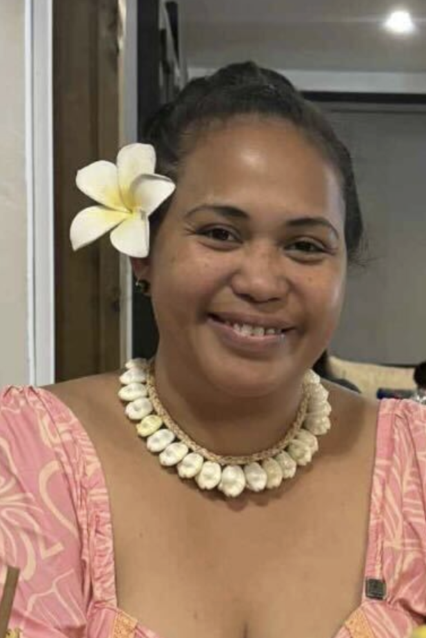 Femme souriante portant un collier de coquillages, une fleur dans les cheveux et une robe rose avec un motif floral, dans un environnement intérieur.
