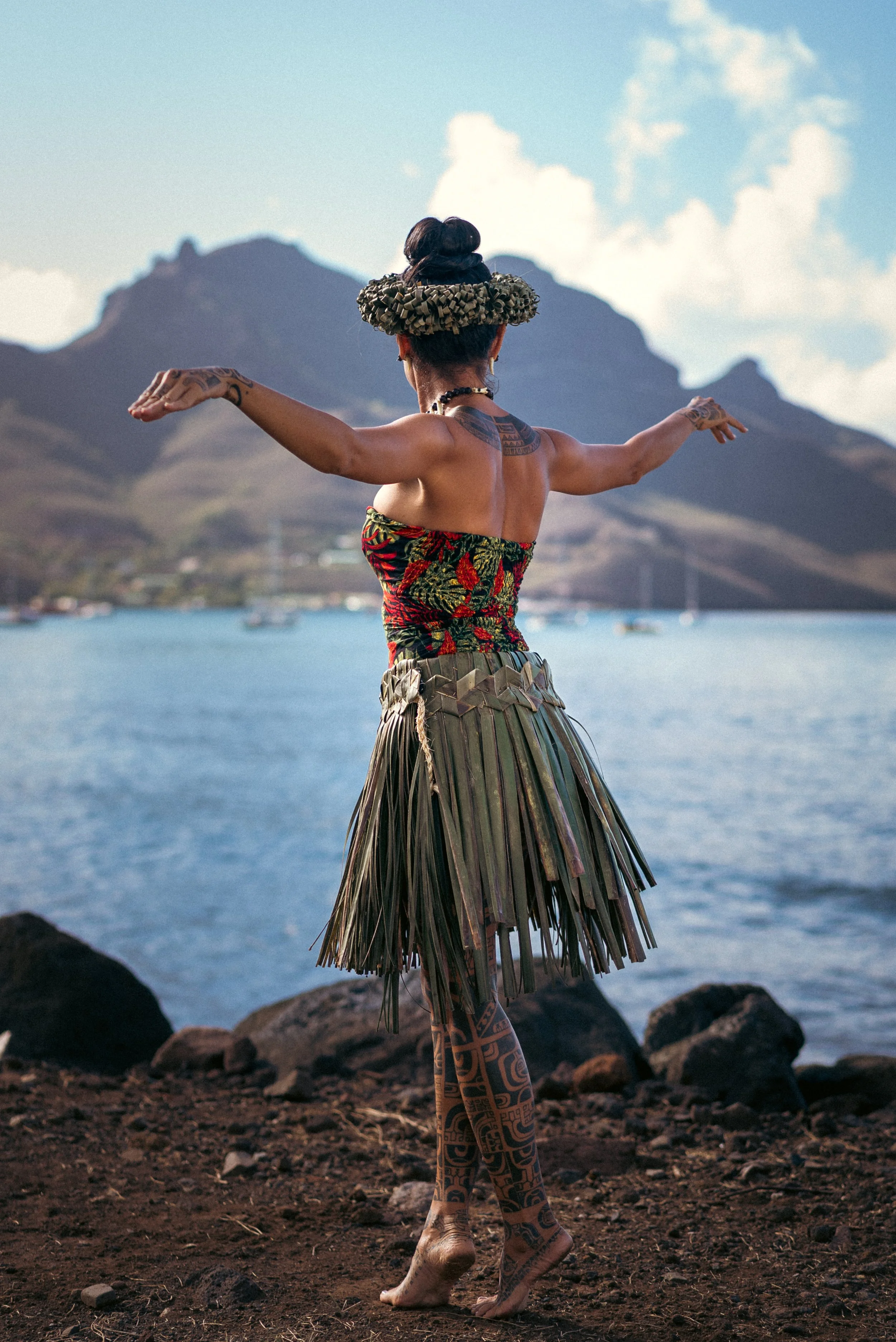 Une femme nue vue de dos avec des tatouages, portant un haut coloré et une jupe en feuilles, en position de danse ou de méditation près de l'eau, avec des montagnes en arrière-plan.