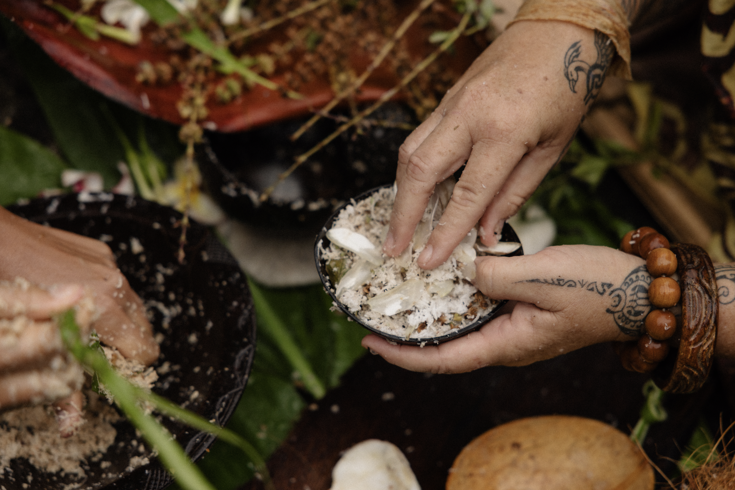 Une personne avec des tatouages, utilisant une cuillère pour probablement servir de la nourriture d'un récipient noir, entourée de plats et d'éléments naturels comme des plantes et de la terre.