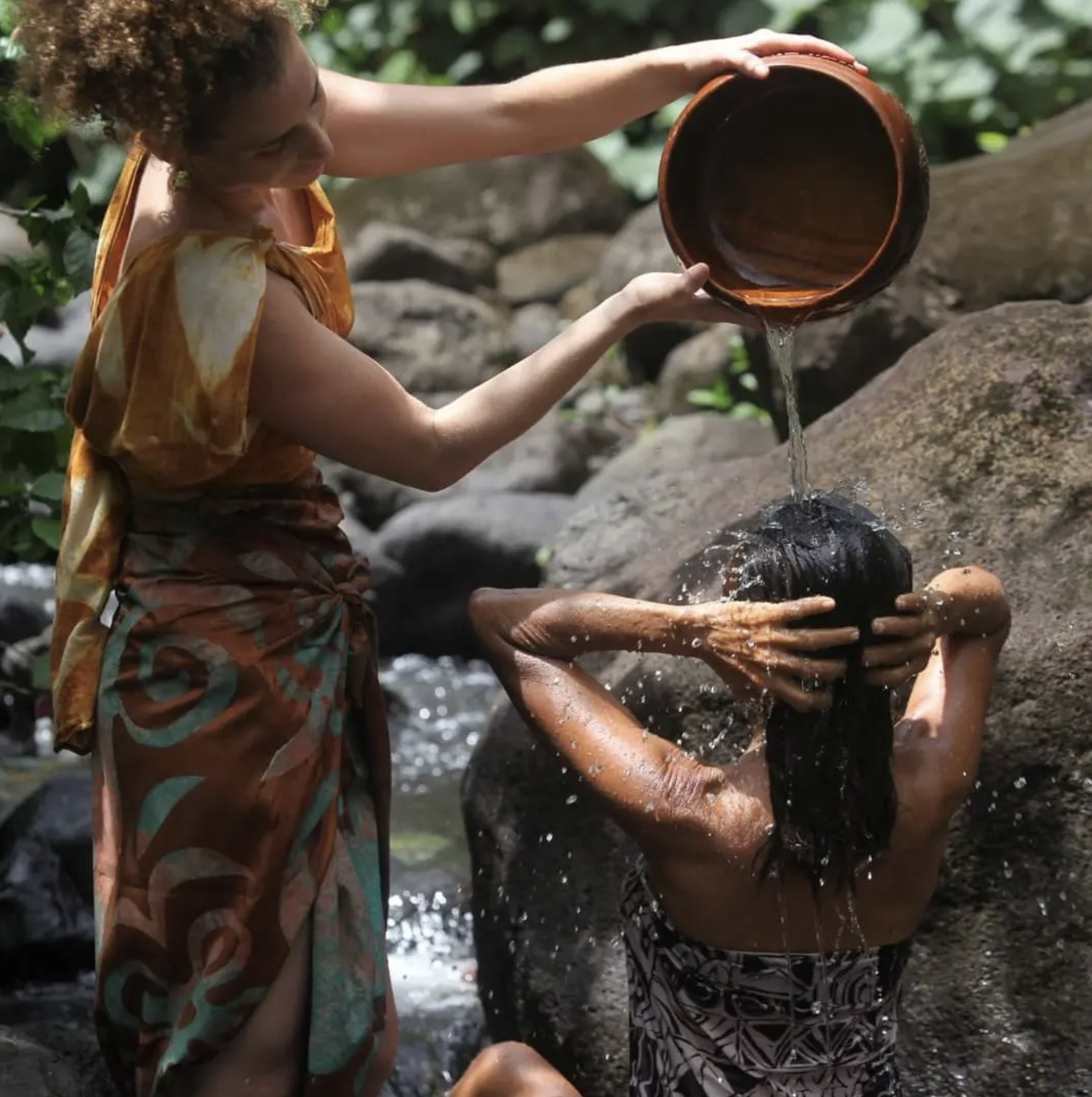Deux femmes, l'une debout avec une écharpe colorée, verse de l'eau d'un pot en terre sur une autre femme qui est abaissée dans une rivière, en train de se laver les cheveux.
