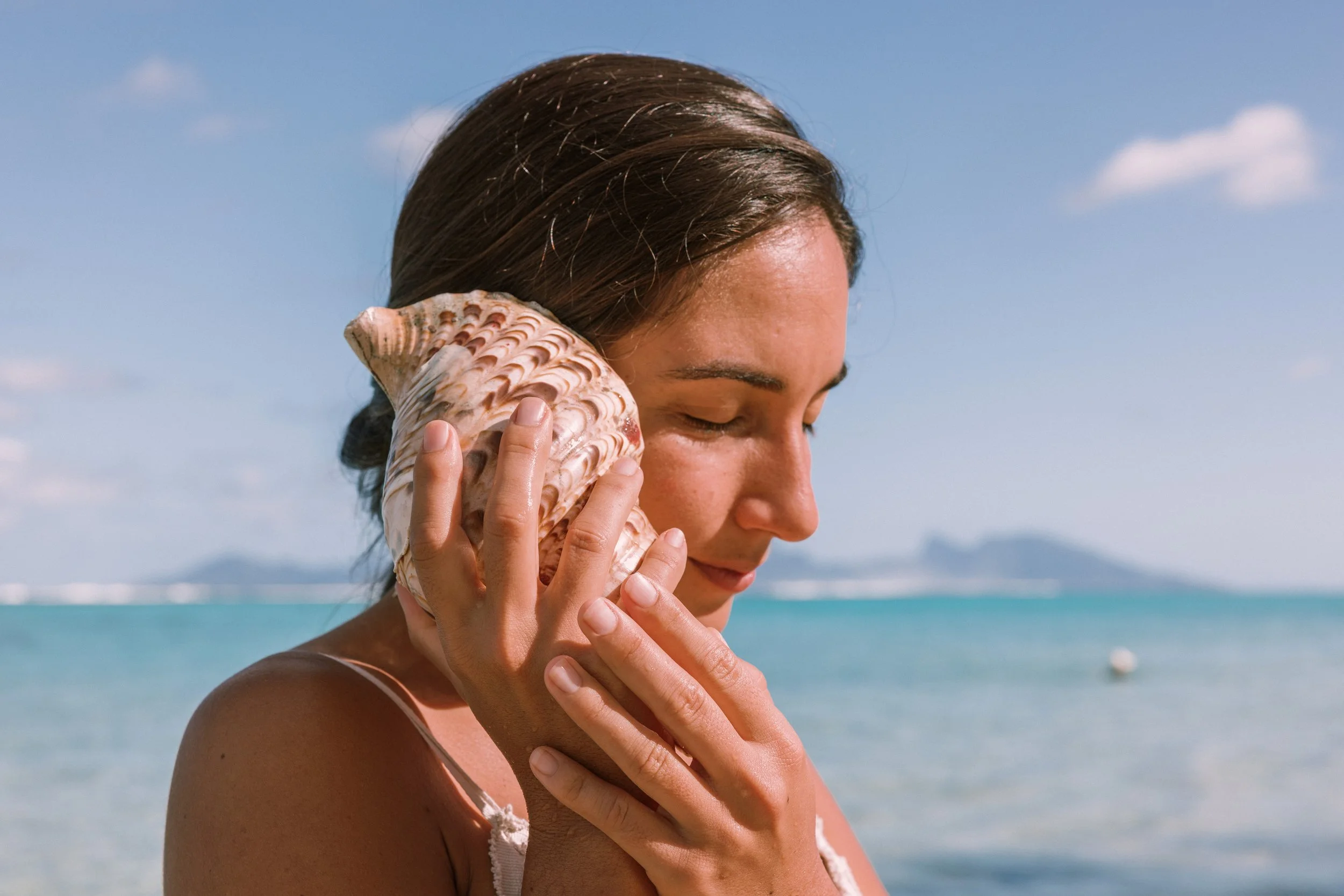 Une femme à la plage avec la mer en arrière-plan, tenant un coquillage près de l'oreille et souriante.