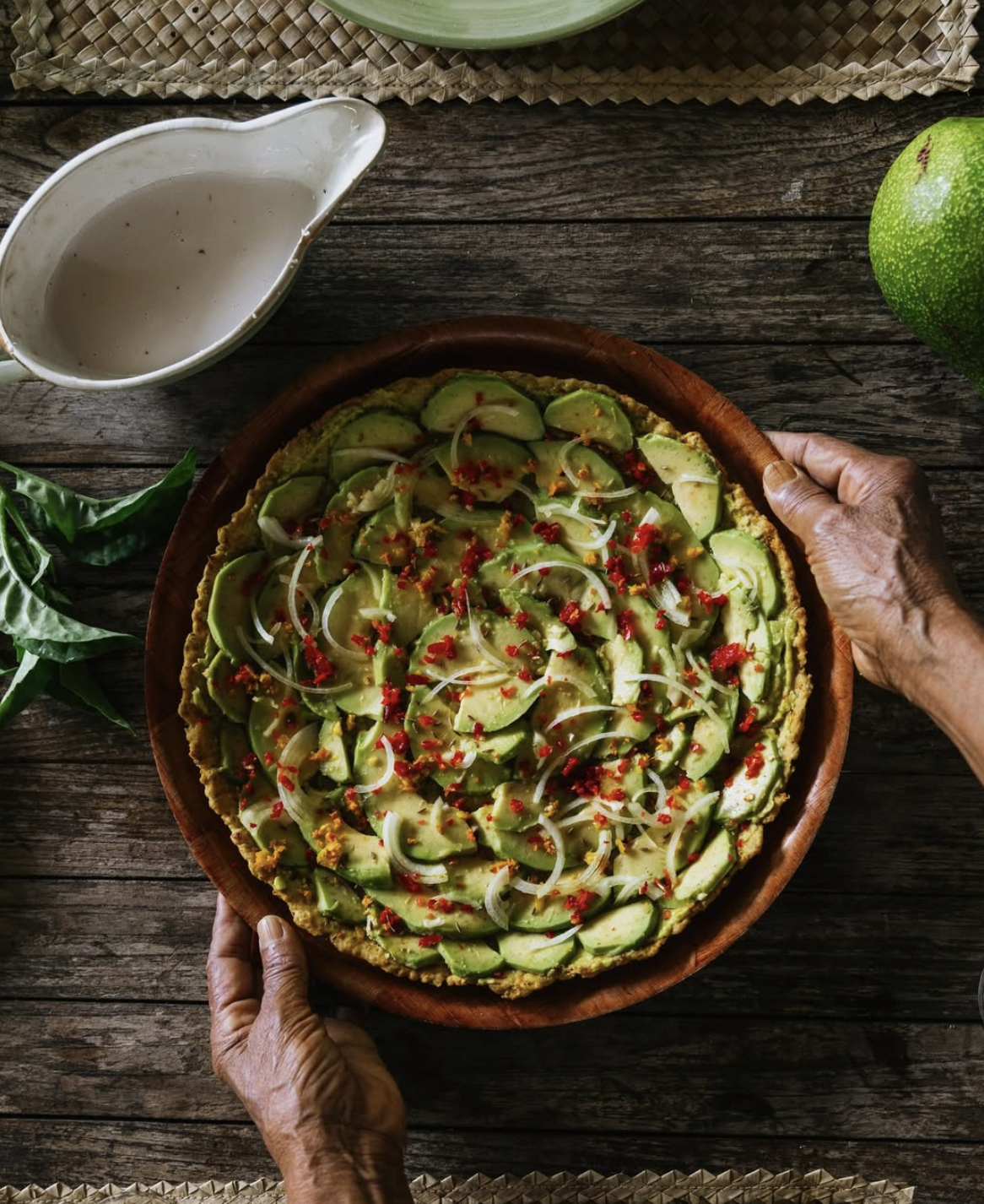 Une personne tenant une pizza végétarienne avec avocat, oignons et épices, sur une table en bois.