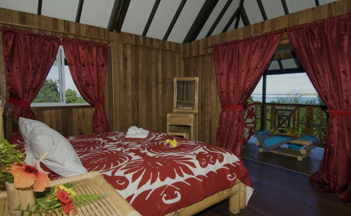 Chambre en bois avec un lit à draps rouges et blancs, deux fenêtres avec rideaux rouges, coin salon extérieur avec mobilier en bois et vue sur la nature.