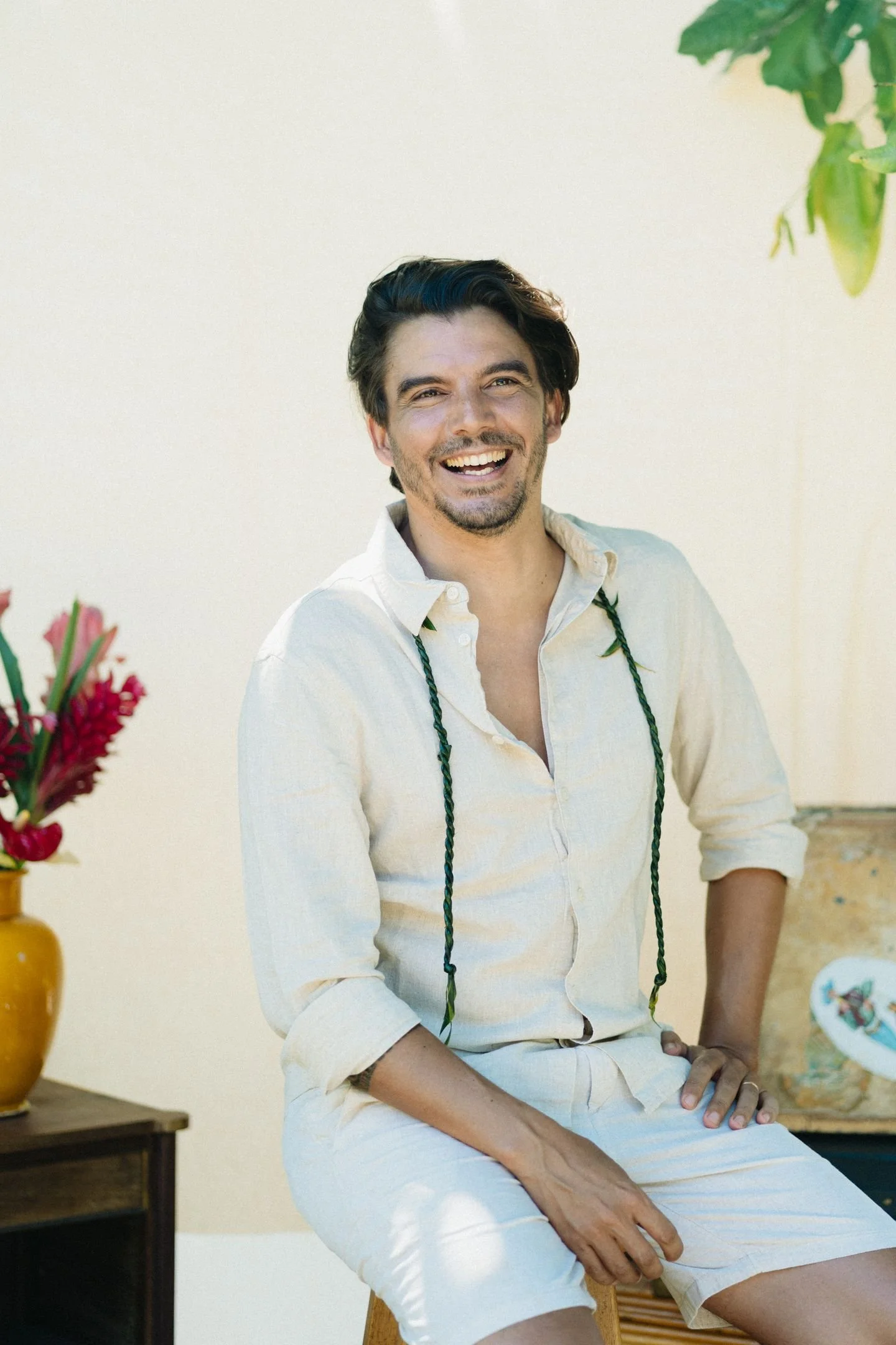 Un homme souriant, habillé d'une chemise beige clair et d'un pantalon beige, assis sur un tabouret en bois, avec une pièce orange à sa droite, des fleurs rouges dans un vase et un décor coloré en arrière-plan.