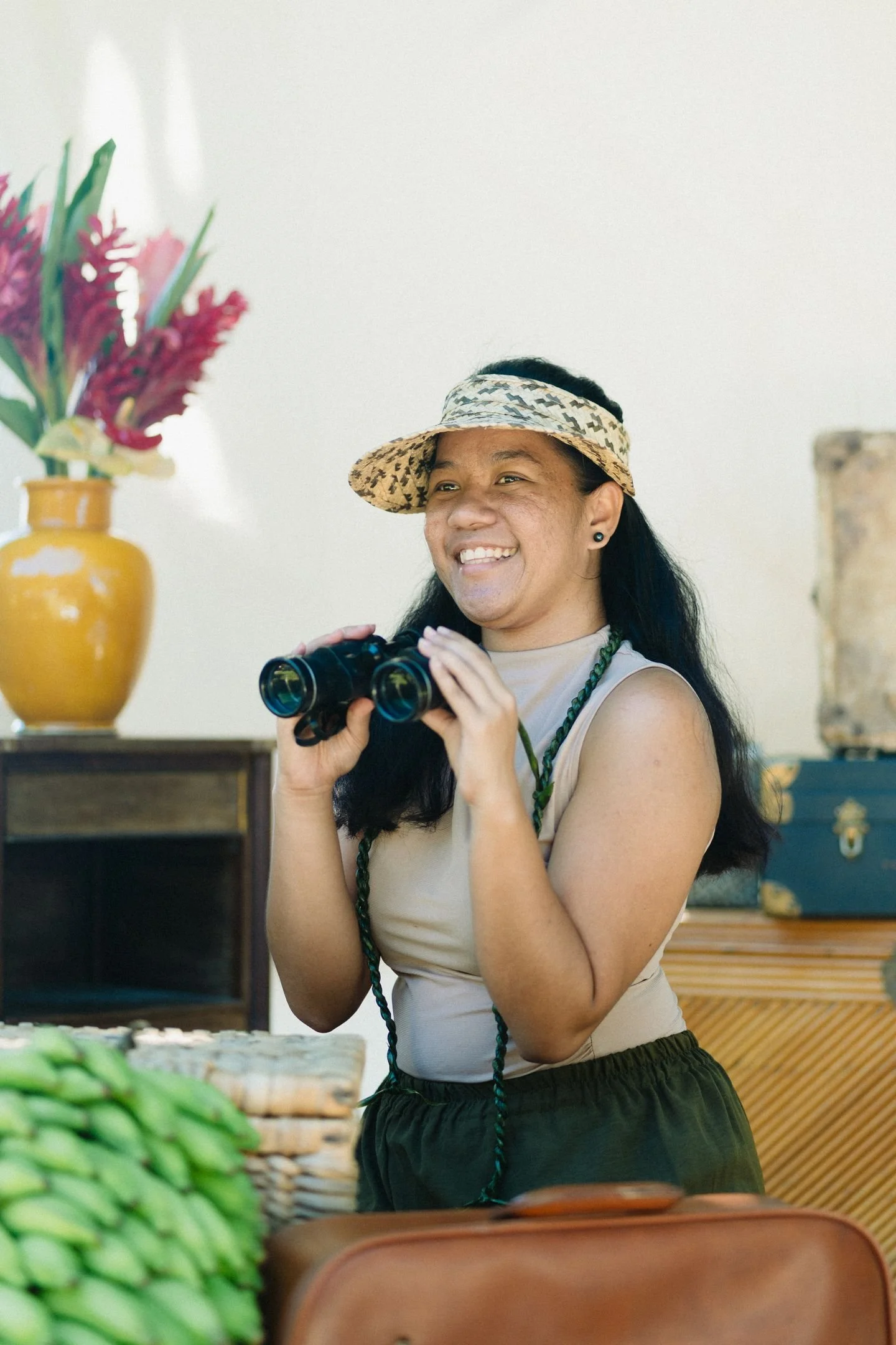 Une femme souriante, portant un chapeau de paille et tenant des jumelles, se tient dans un intérieur avec un décor naturel et végétal.