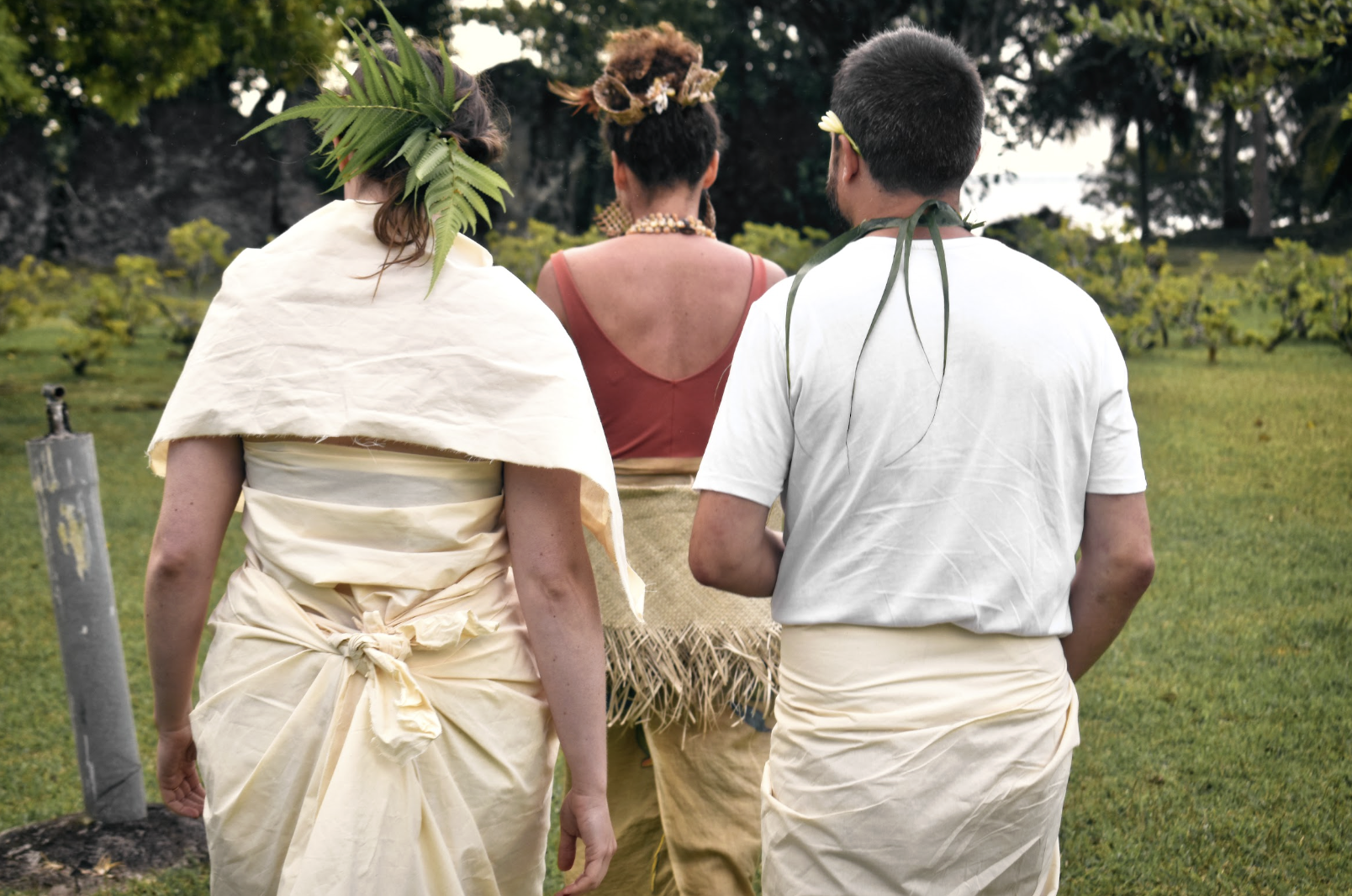 Trois personnes portant des vêtements traditionnels marchent dans un champ verdoyant, avec des arbres en arrière-plan.