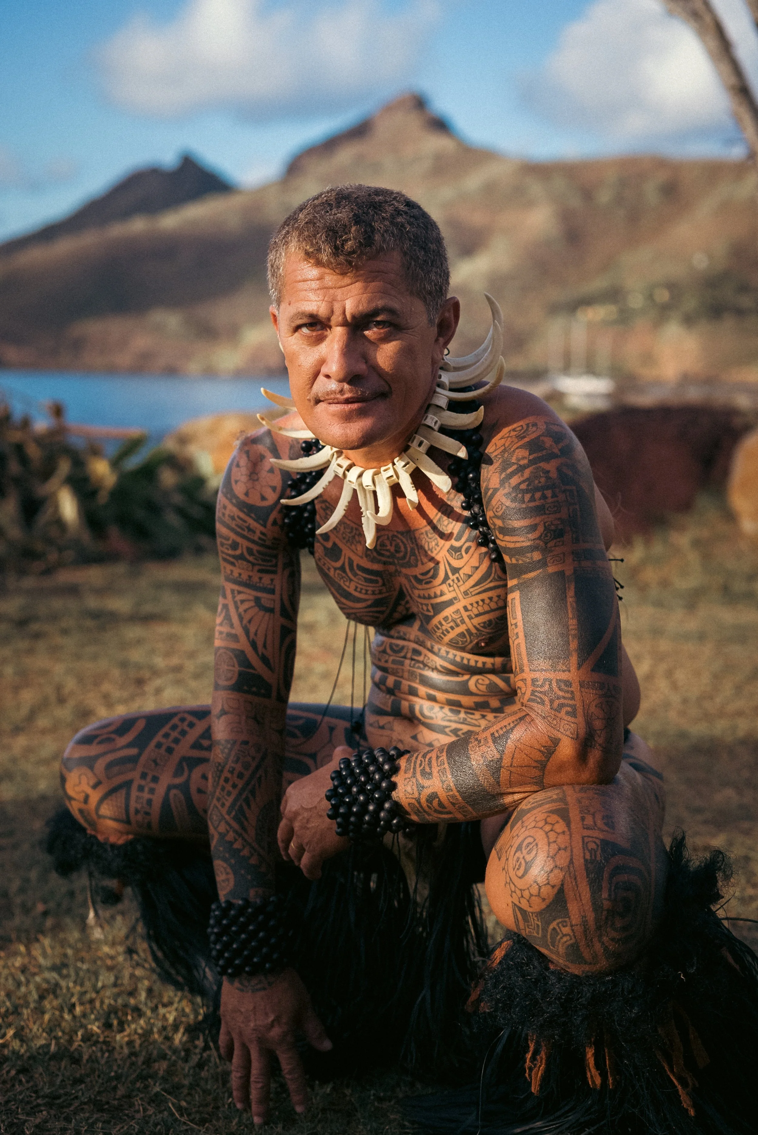 Un homme tatoué et vêtu de vêtements traditionnels maoris, portant un collier en dents ou en os, squat dans un paysage naturel avec une montagne en arrière-plan et un bateau en mer.