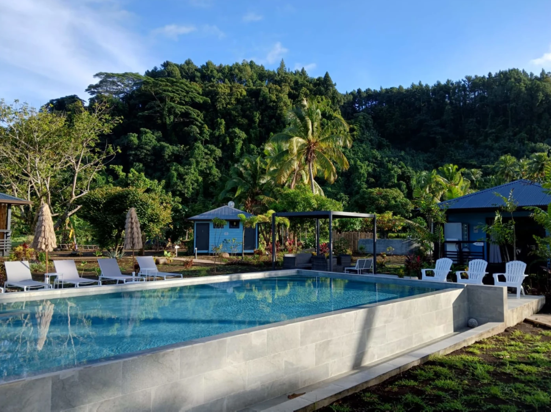 Piscine extérieure dans un cadre tropical avec des chaises longues, des parasols et des petites maisons en toile de fond, entourée de végétation luxuriante et une montagne verte en arrière-plan.