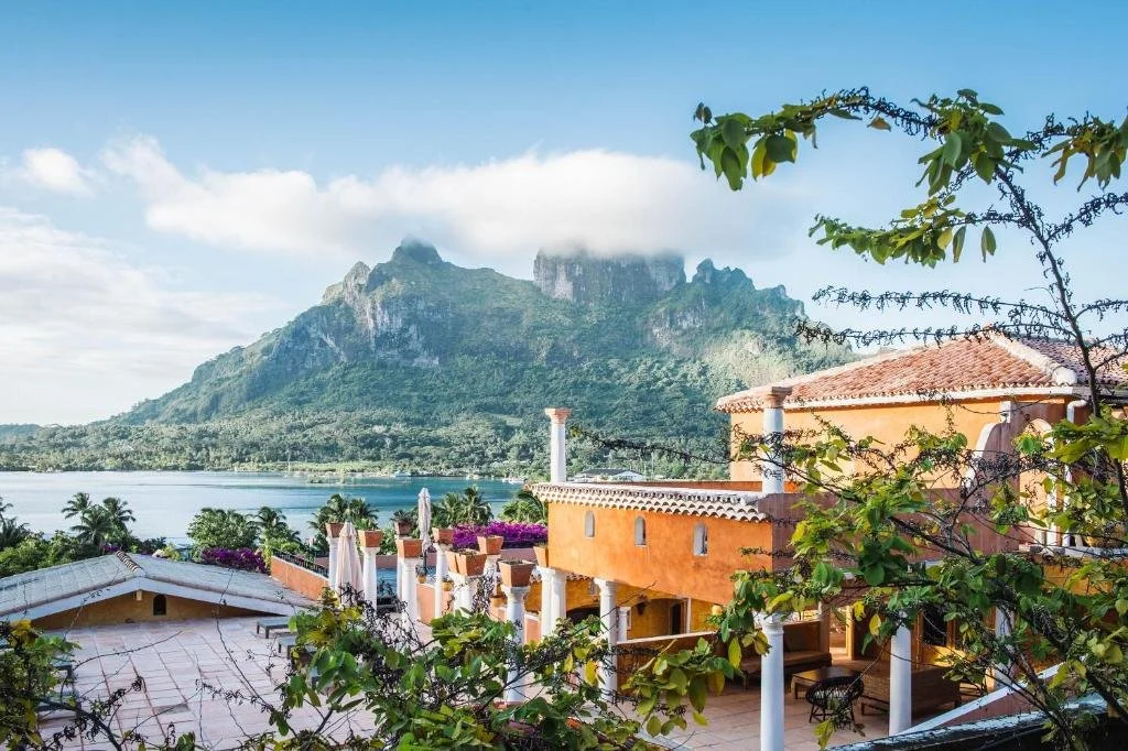 Vue d'une maison méditerranéenne orange avec terrasse, face à un lac entouré de montagnes et de palmiers, sous un ciel partiellement nuageux.