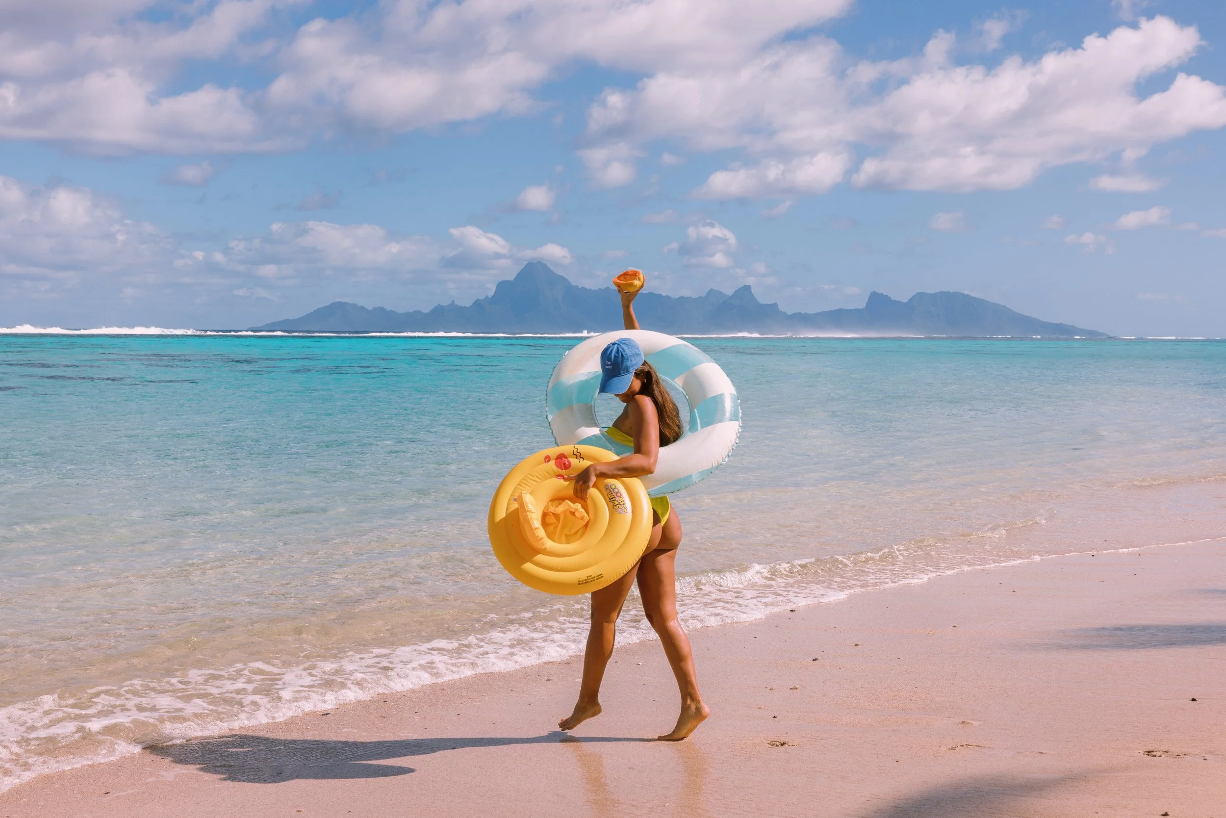 Une femme sur la plage avec un anneau gonflable, portant un chapeau bleu, une serviette de plage jaune, et se tenant au bord de l'eau avec un paysage de montagnes en arrière-plan.