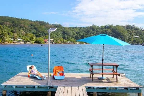 Une personne allongée sur une chaise longue sur un ponton en bois au bord d'un lac, avec un parasol bleu, une table, des chaises, et un paysage vert en arrière-plan.