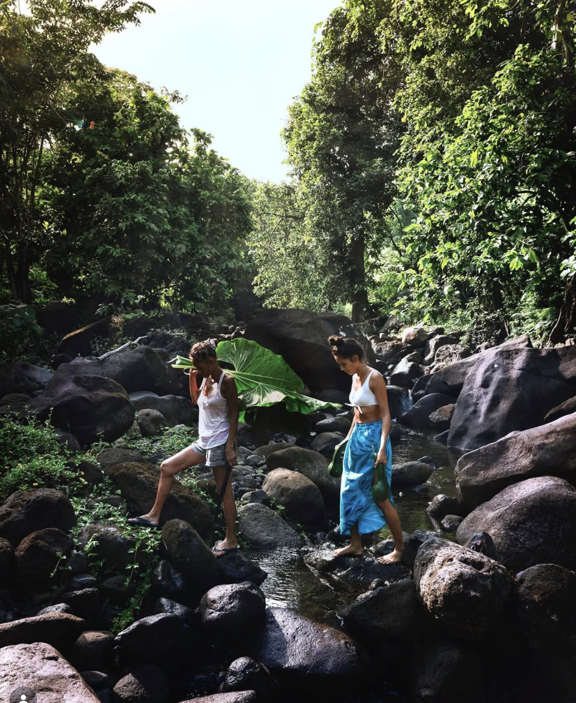Deux femmes traversent un ruisseau dans une forêt dense, l'une portant un grand feuille verte comme parapluie, l'autre avec un sac en plastique vert.