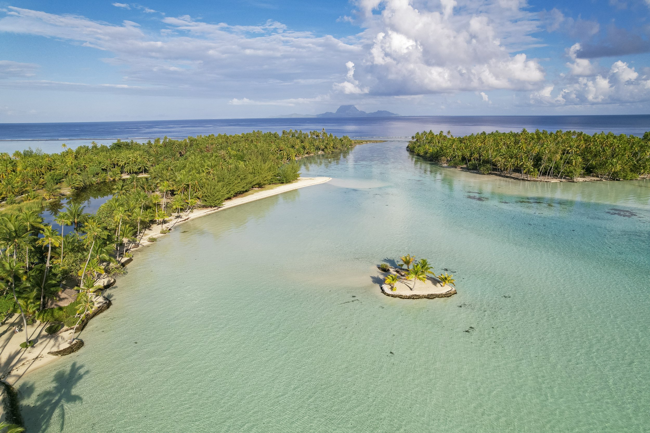 Vue aérienne d'une lagune tropicale bordée de palmiers, avec l'océan et une île lointaine en arrière-plan, ciel partiellement nuageux.