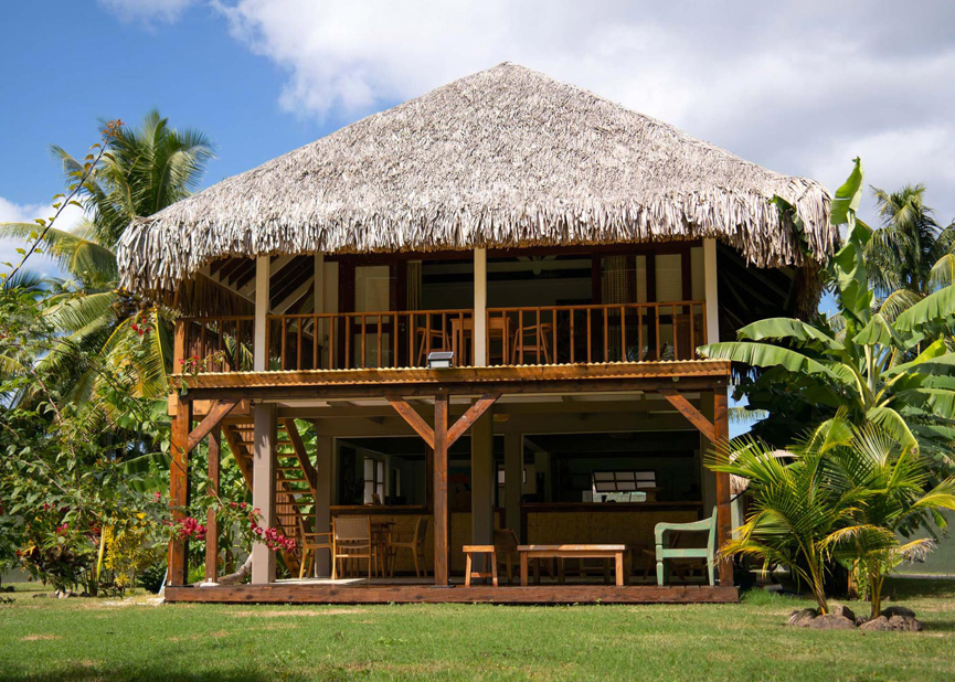 Une villa en bois avec un toit en chaume, entourée de végétation tropicale, sous un ciel partiellement nuageux.