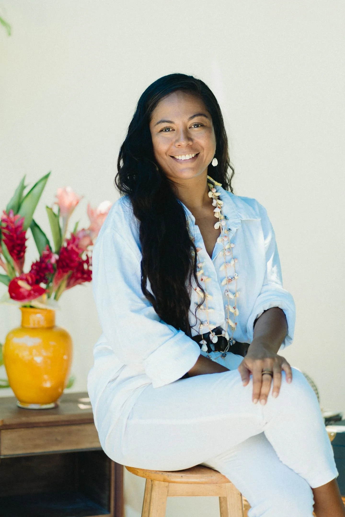 Une femme souriante avec des cheveux longs noirs, portant une chemise blanche et un collier de coquillages, assise sur un tabouret en bois, à côté d'une table avec un vase jaune contenant des fleurs tropicales. Fond blanc.