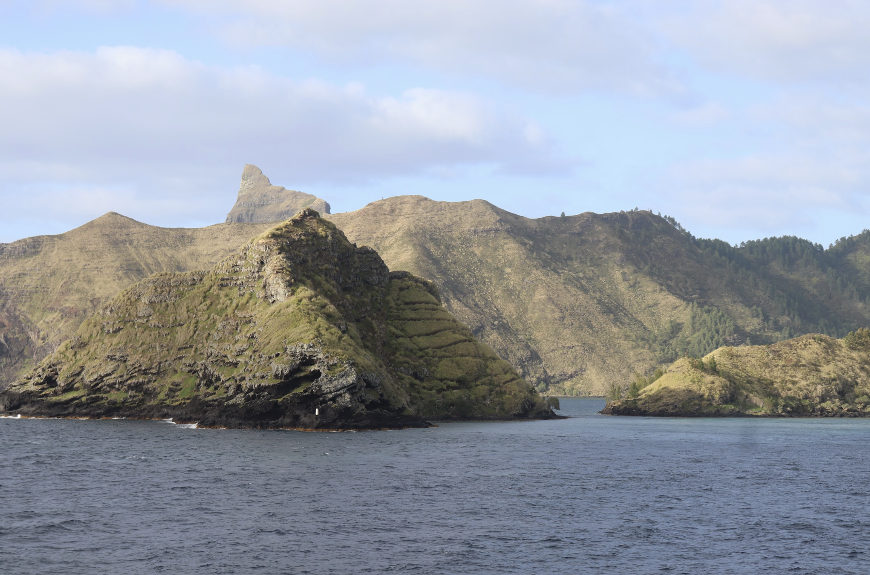 Paysage maritime avec des montagnes verdoyantes et une mer au premier plan, ciel partiellement nuageux.