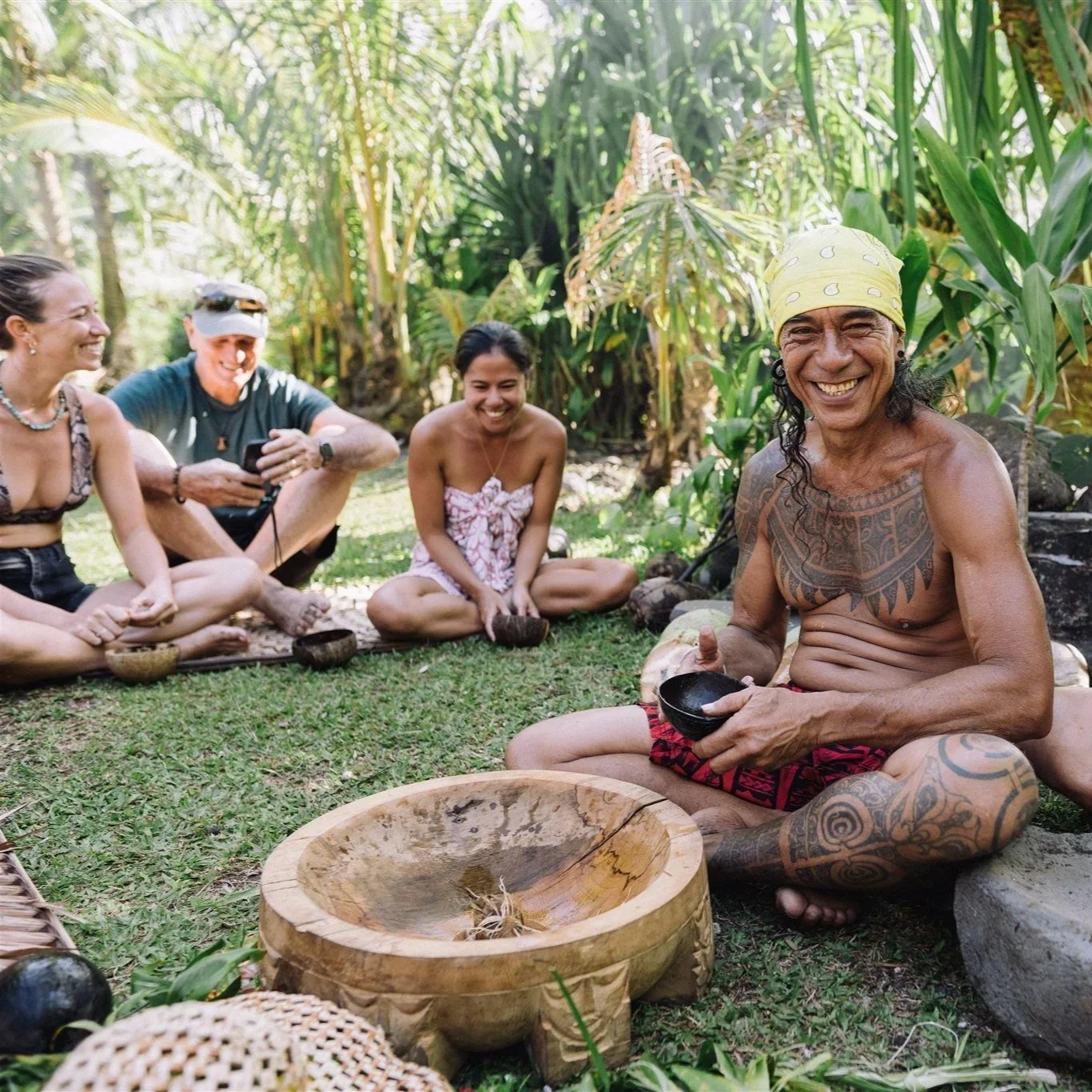 Un groupe de personnes souriantes, y compris un homme indigène tatoué portant un bandana jaune, assis sur l'herbe dans une forêt tropicale, participant à une activité culturelle ou cérémoniale.