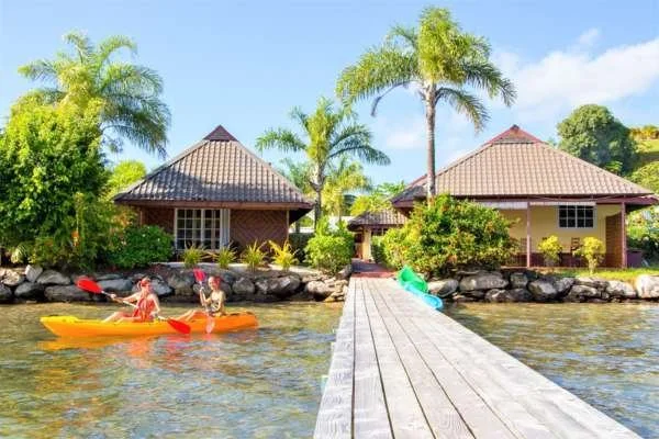 Deux personnes paddellent un kayak orange sur une rivière près de maisons entourées de palmiers et de végétation tropicale.
